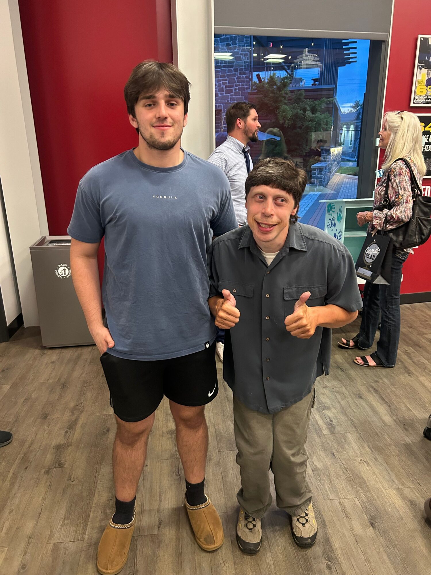 Two men standing indoors, one giving a thumbs-up, the other with hands in pockets, with a window and people in background.