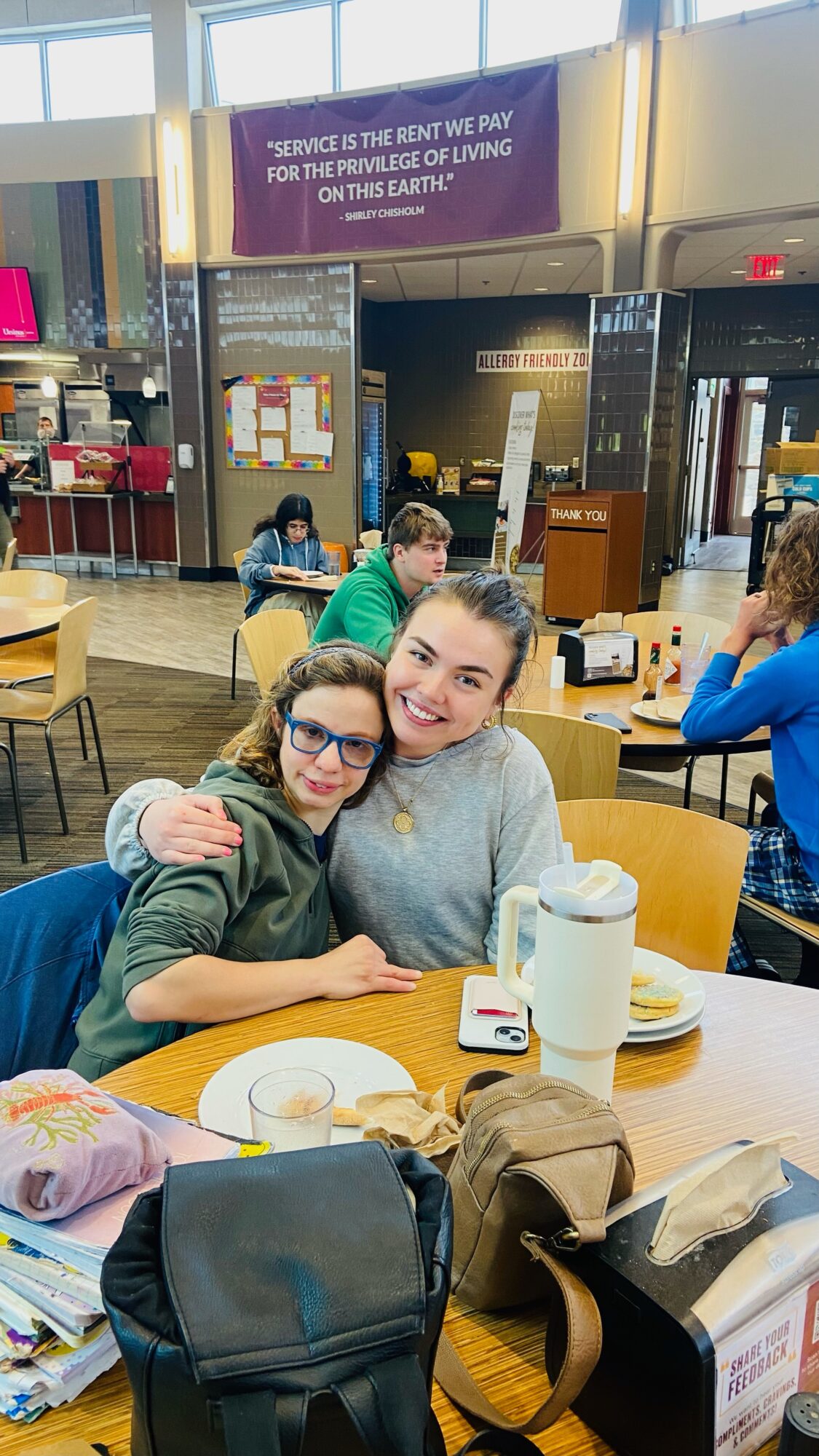 Two smiling women sit at a table in a busy indoor space, with other people and tables visible in the background.