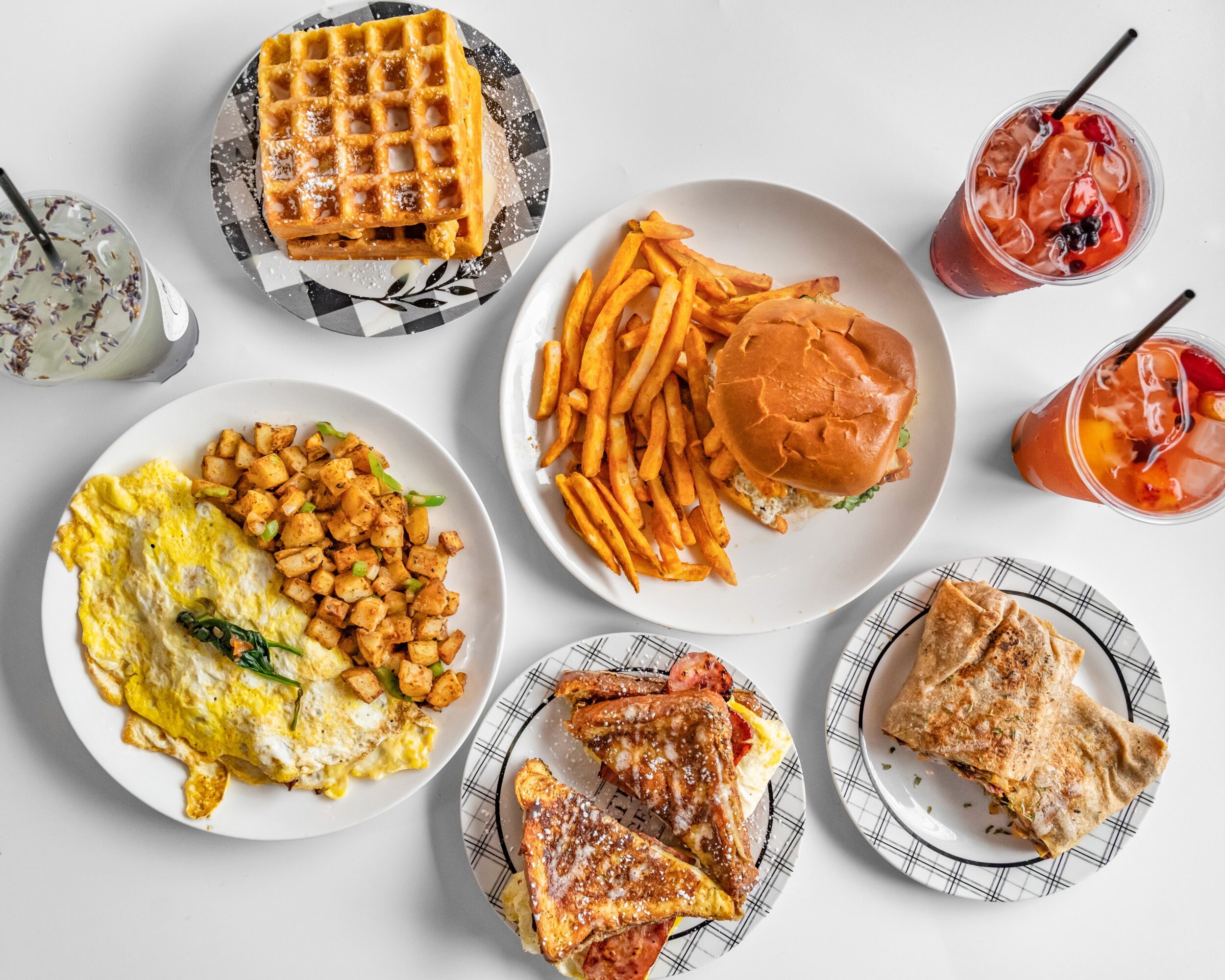 Assorted breakfast foods including waffles, fries with burger, scrambled eggs with chickpeas, toast, and drinks on a white table.