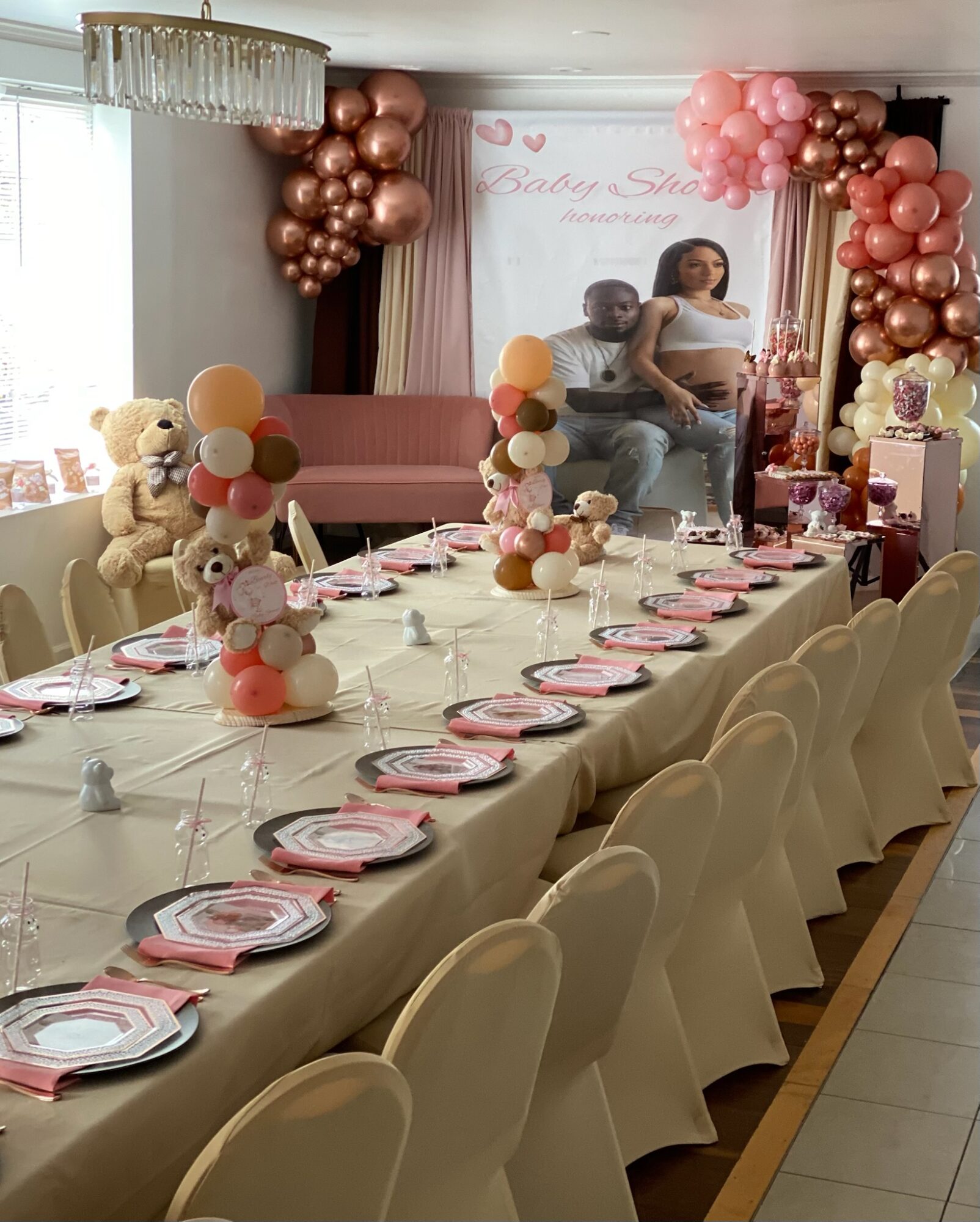 Decorated room with a long table set for a celebration, pink and peach balloons, a teddy bear, and a backdrop with people.