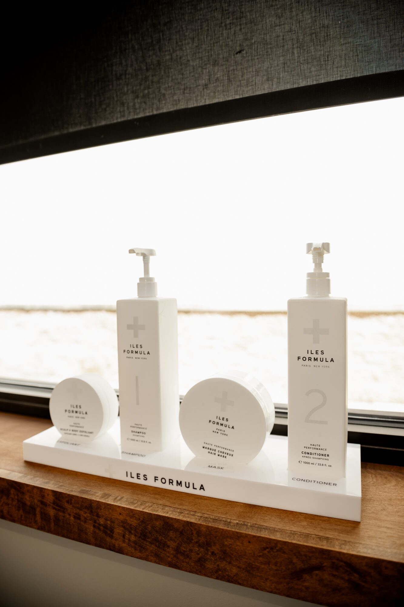Two bottles and two round containers of skincare products on a windowsill with a bright outdoor view.