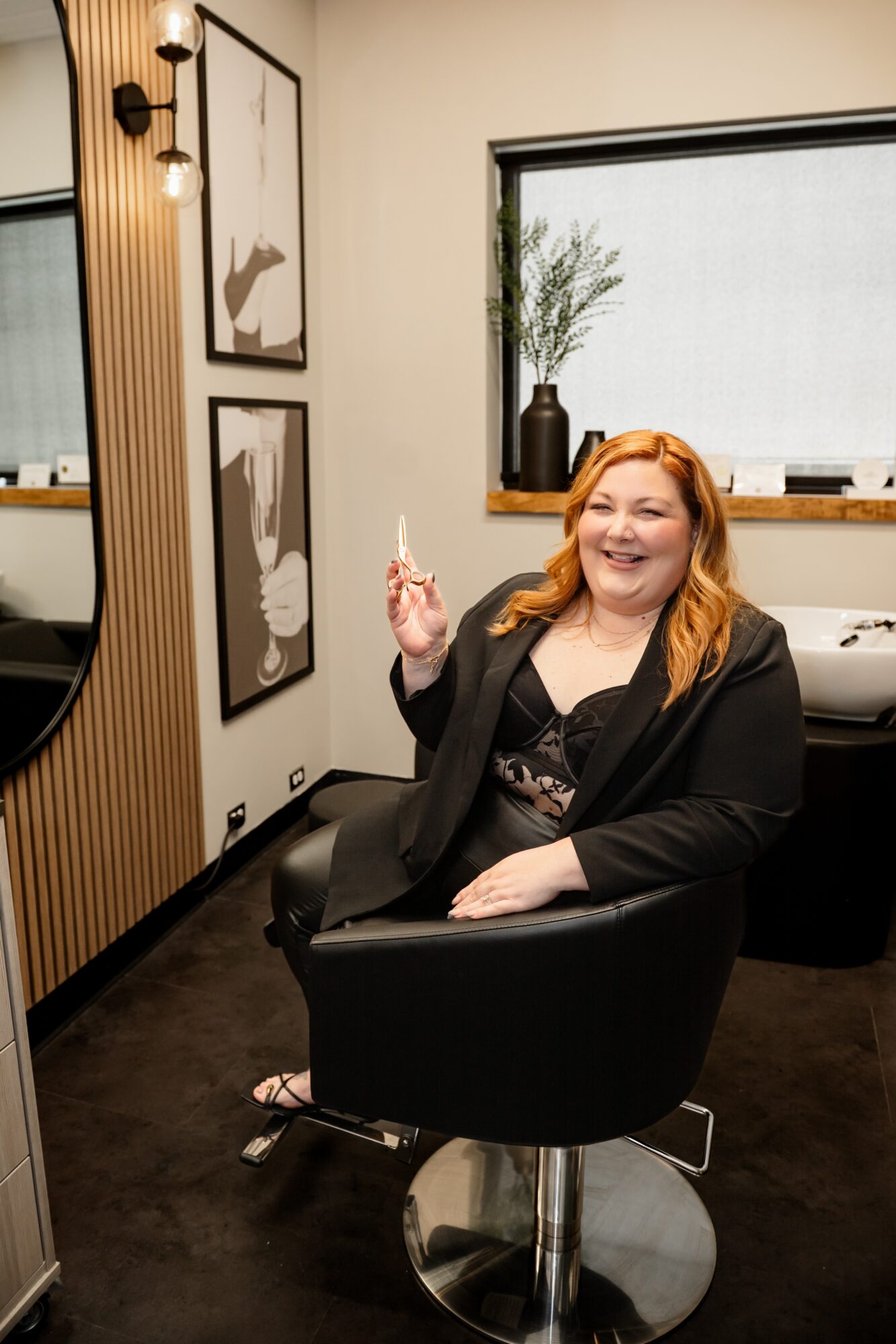 A woman with red hair sitting in a black salon chair, smiling, holding a small object, in a modern interior with artwork and a window.