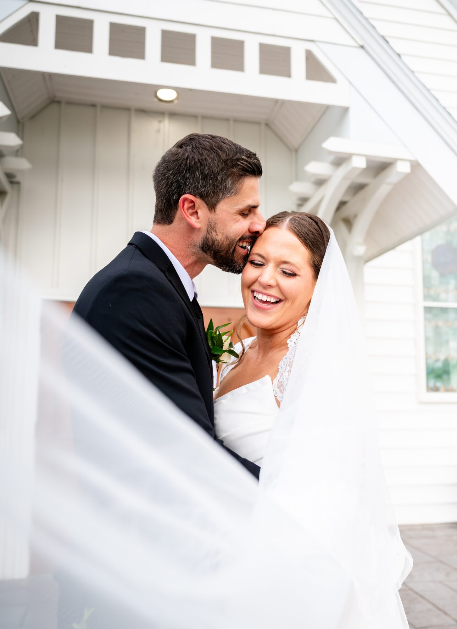 A smiling bride and groom embrace indoors, with the groom whispering into the bride's ear, white wedding dress and suit, bright background.