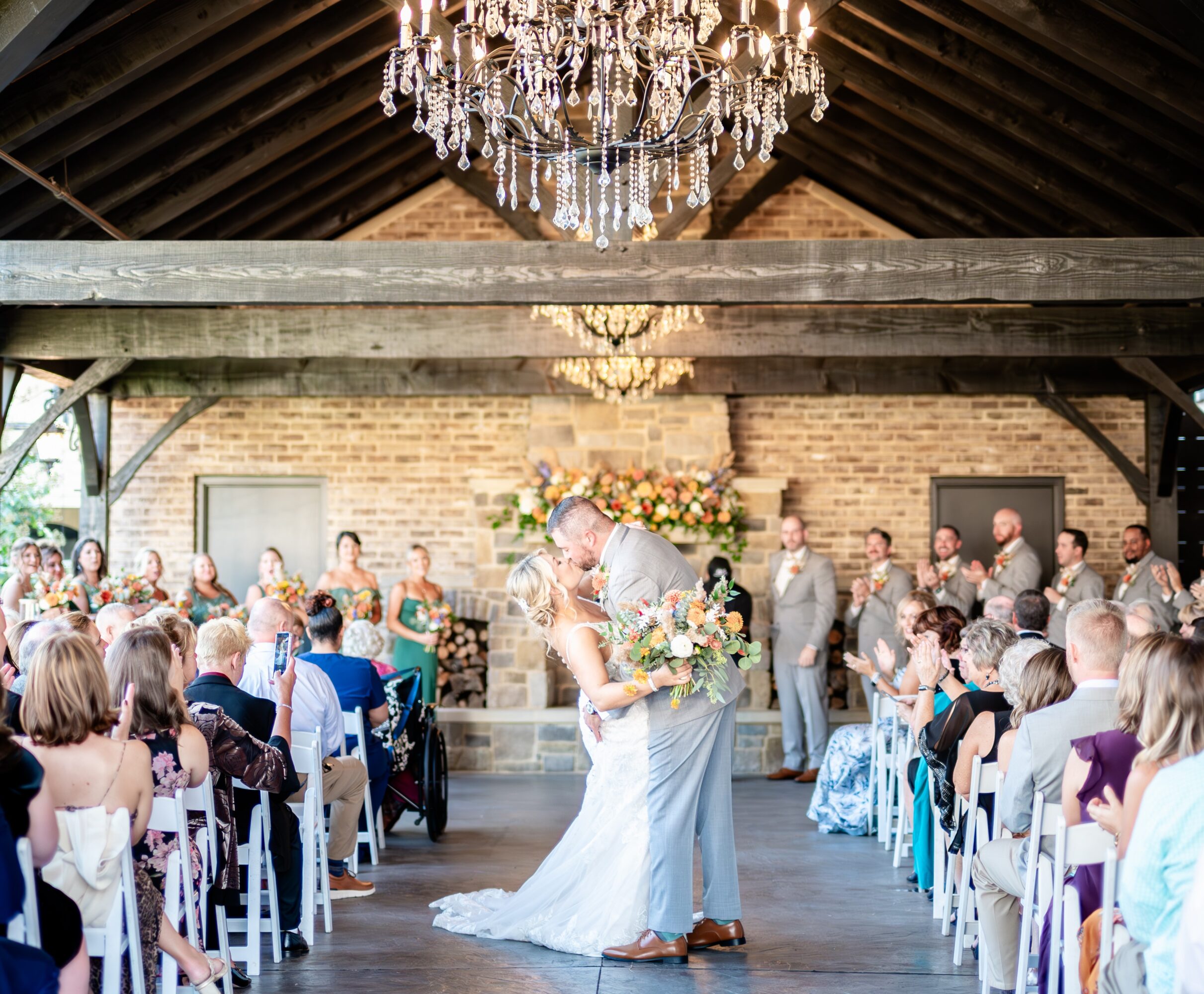 Bride and groom kissing under a chandelier in a rustic wedding venue with guests seated on both sides.