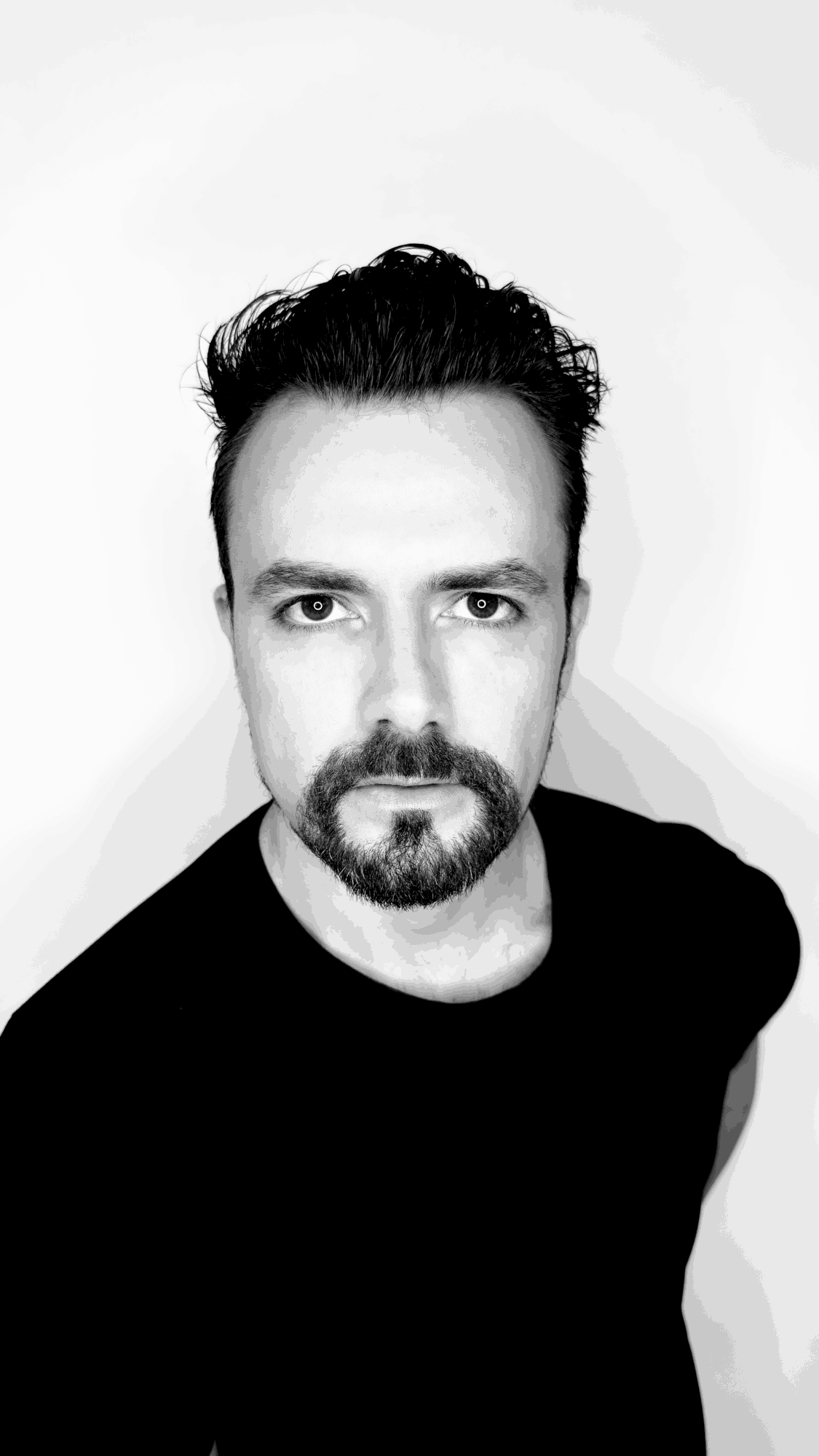 Black and white portrait of a man with dark hair and beard, wearing a black shirt, looking directly at the camera.