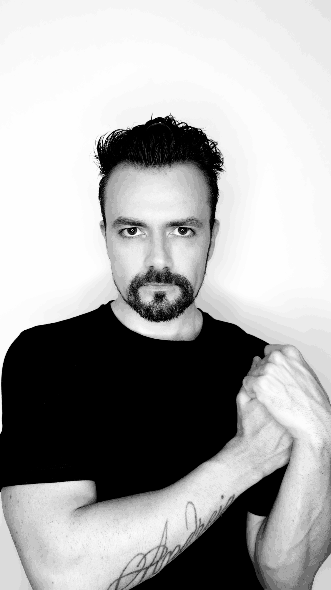 Man with dark hair and beard, wearing a black shirt, holding his arm, against a plain white background.