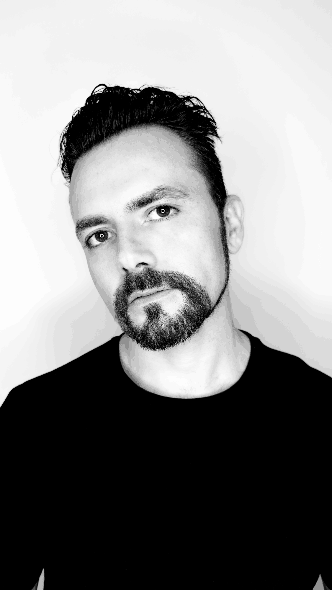 Portrait of a man with dark hair and a beard, looking at the camera, wearing a black shirt, against a plain background.
