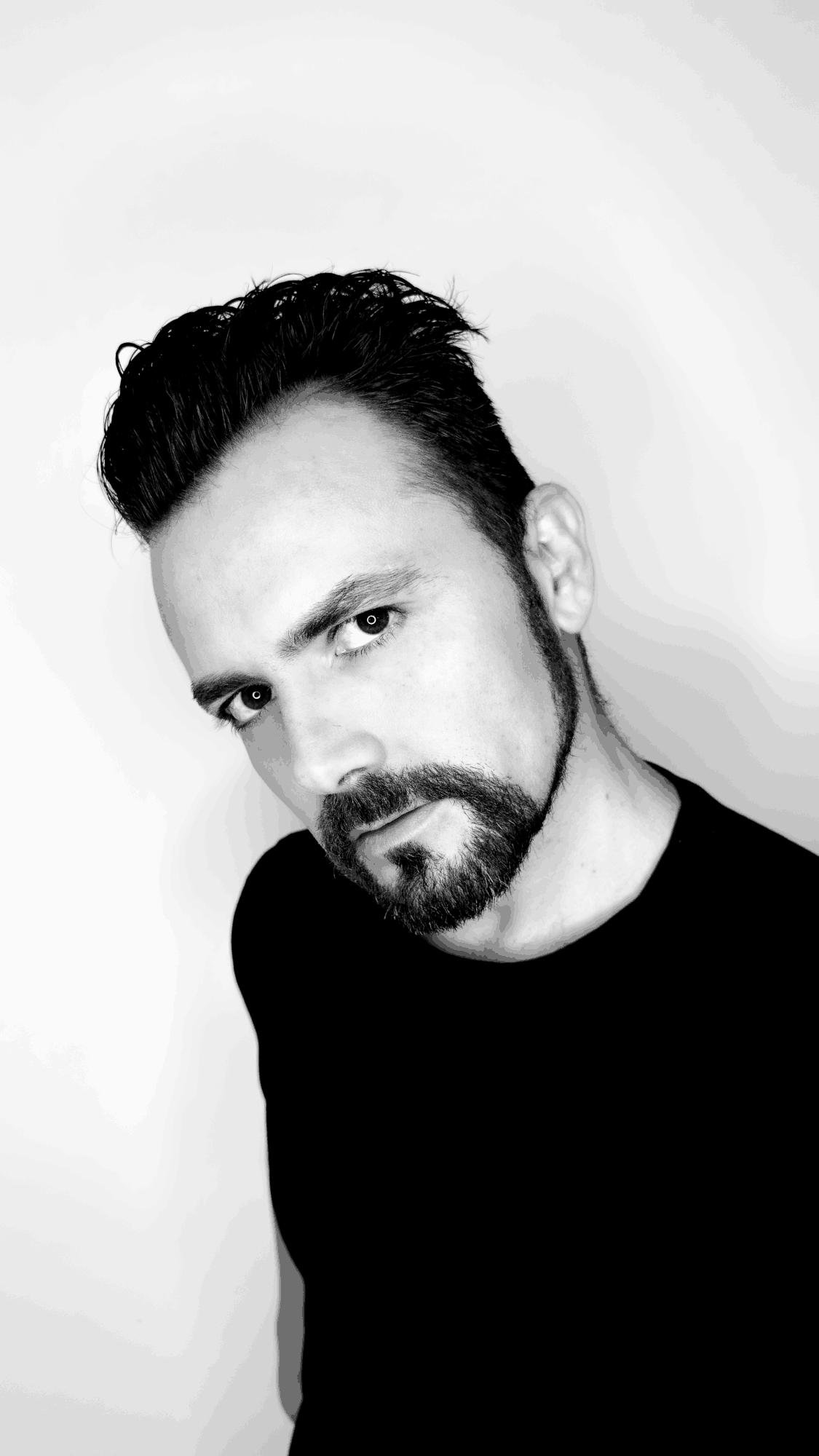 Man with dark hair and beard looking at camera, head tilted, black shirt, plain background.