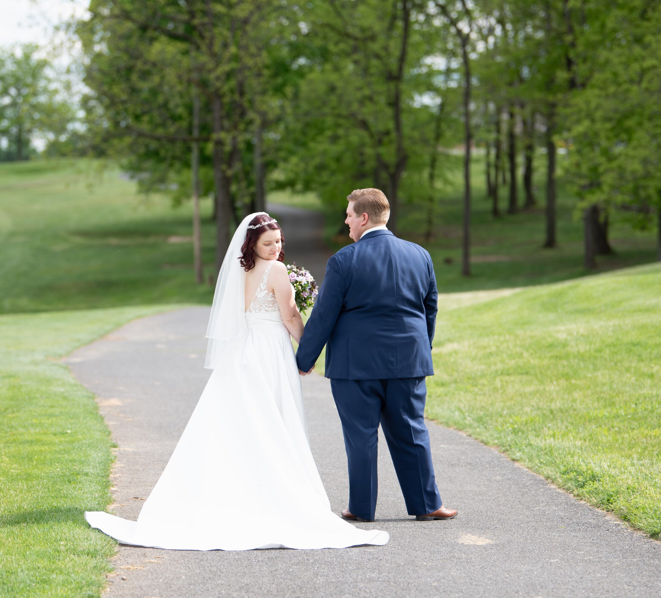 Bride and groom holding hands on a park path, surrounded by green trees and grass.