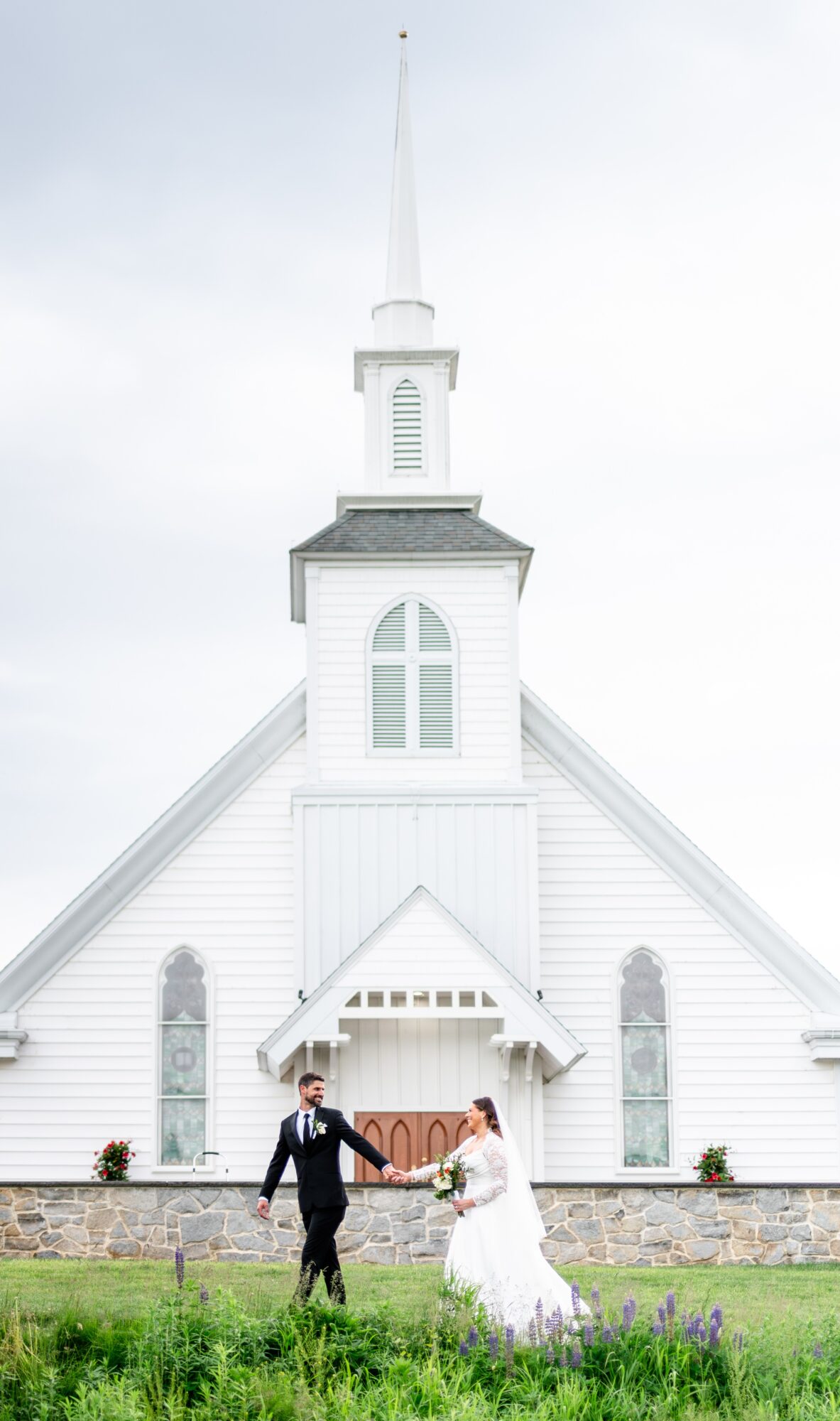 A couple in wedding attire holding hands in front of a white church with a tall steeple, grass and flowers in foreground.
