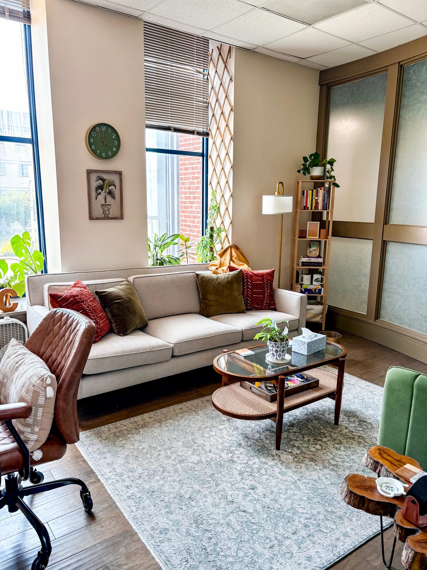 Living room with white sofa, wooden coffee table, green and red cushions, large windows, bookshelf, and potted plants.