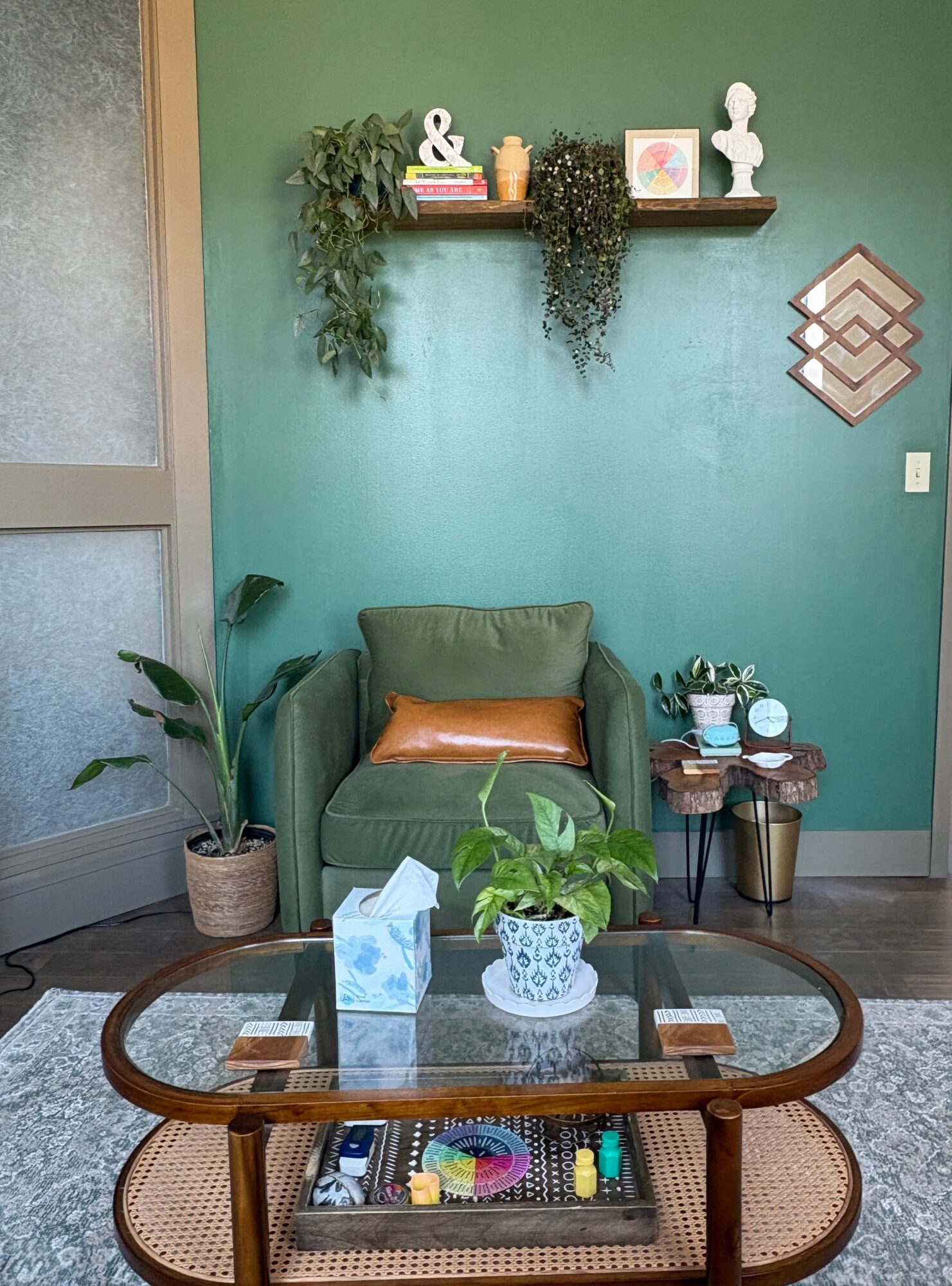 Green armchair with a brown pillow, small table with plants and books, wall shelf with decorative items, and a patterned wall decor.