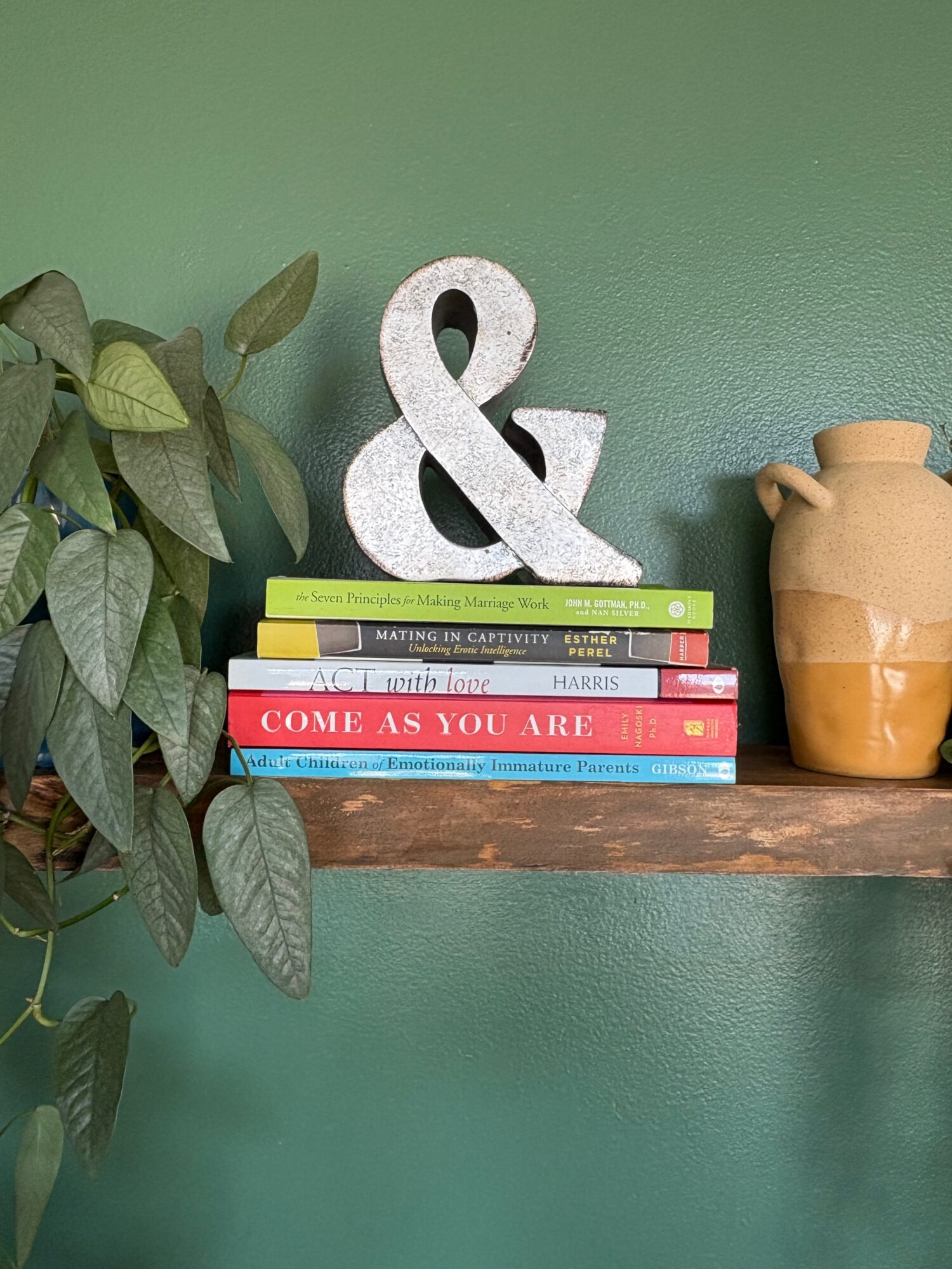 Shelf with books, a decorative ampersand, a potted plant, and a vase against a green wall.