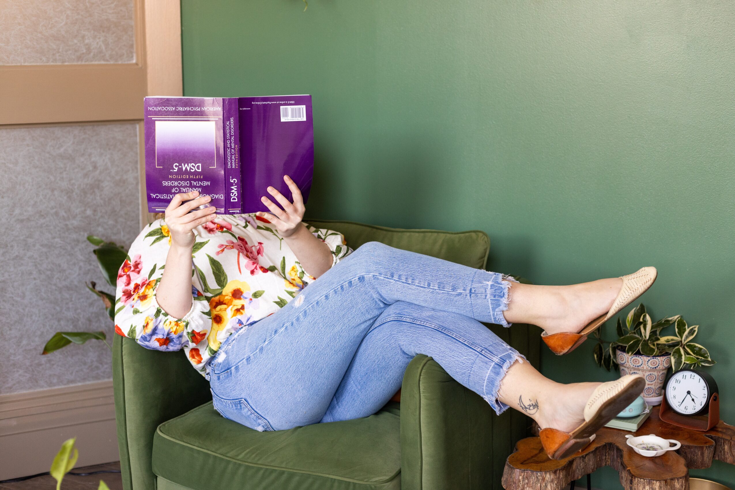 Person lying on green sofa with legs crossed, holding a purple book in front of face, wearing floral blouse and jeans.