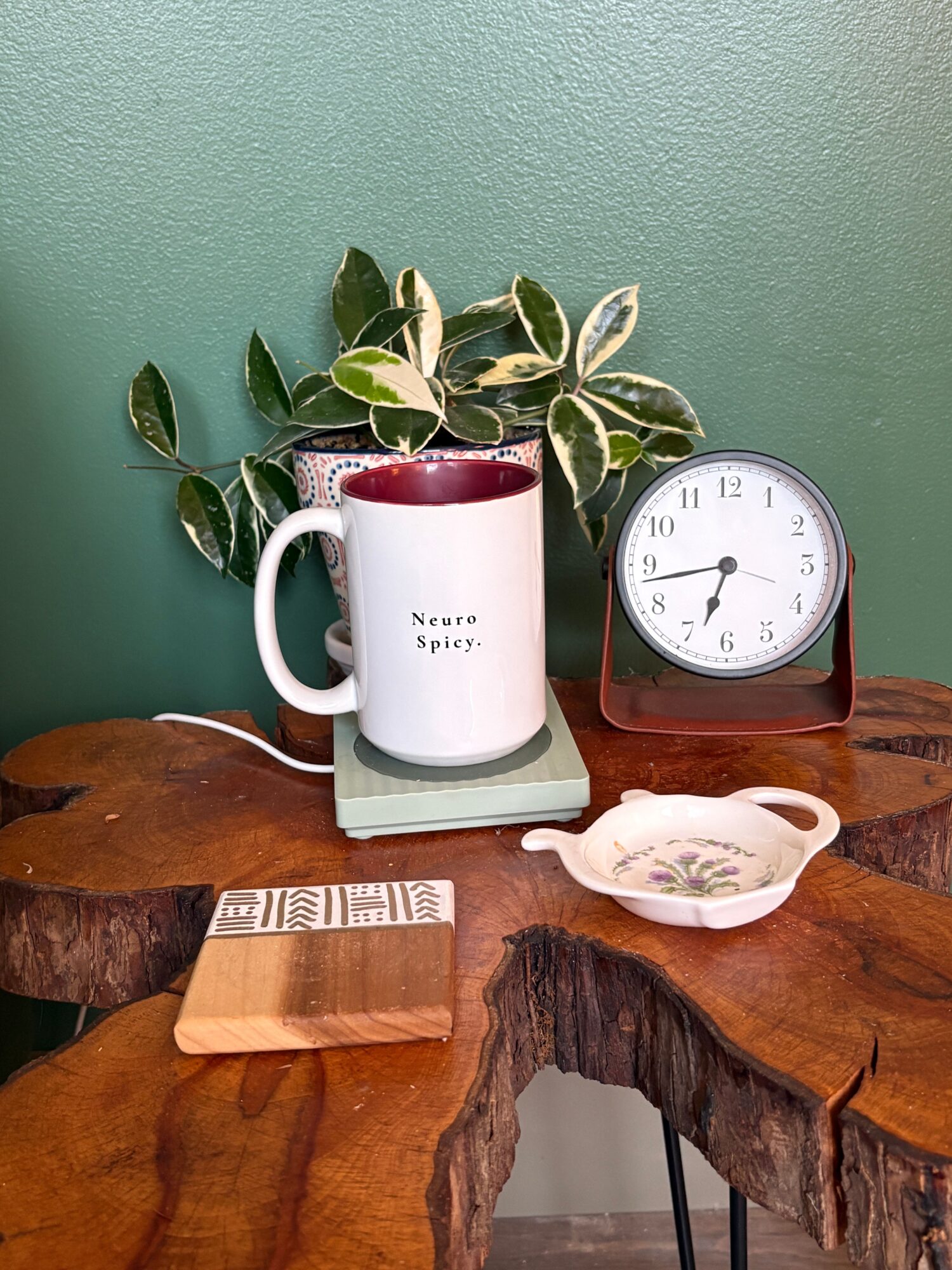 Clock, plant, mug, teacup, and wooden block on a wooden surface against a green wall.