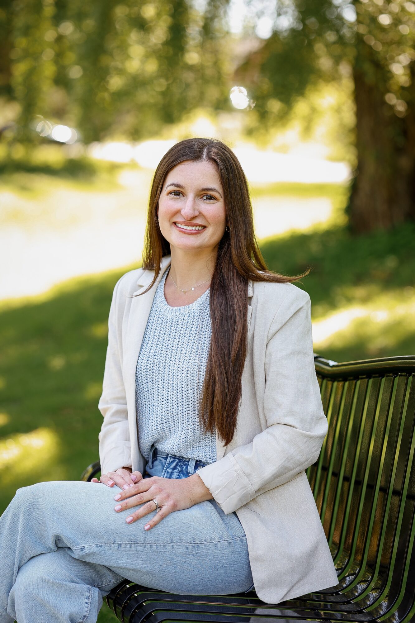 Young woman with long brown hair smiling, sitting on a park bench in sunlight, wearing a light blazer and jeans.