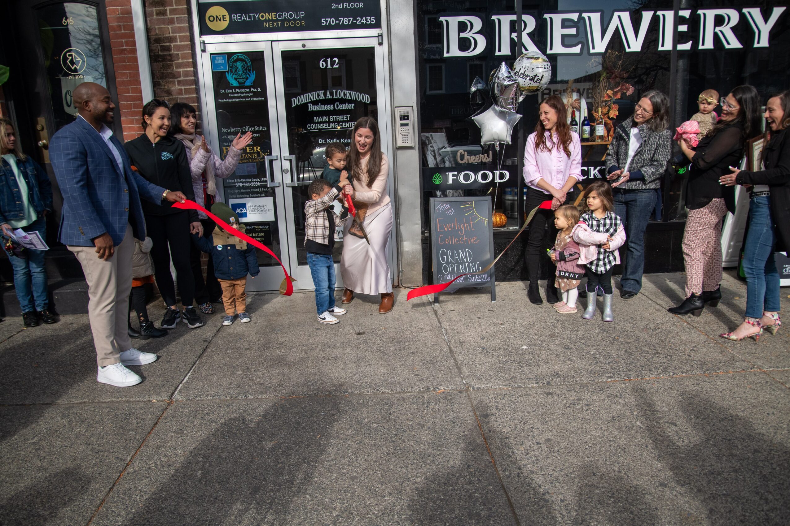 Group of people gathered outside a brewery, some holding balloons, children present, on a city sidewalk, daytime.