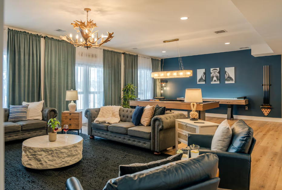 Living room with sofas, a round coffee table, curtains, and wall art, illuminated by a chandelier and natural light.