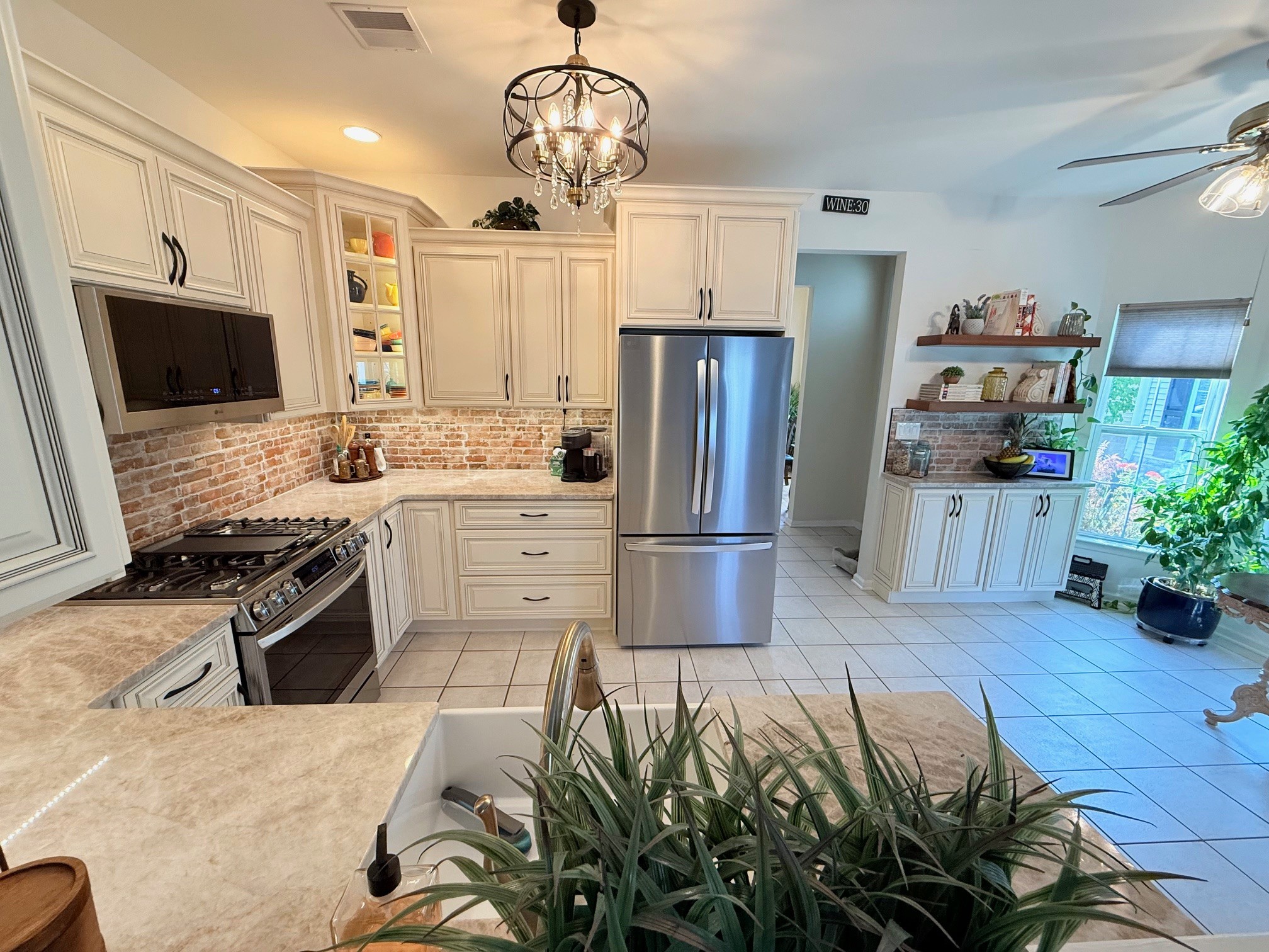 Kitchen with white cabinets, stainless steel refrigerator, brick backsplash, and a chandelier, with a plant in the foreground.
