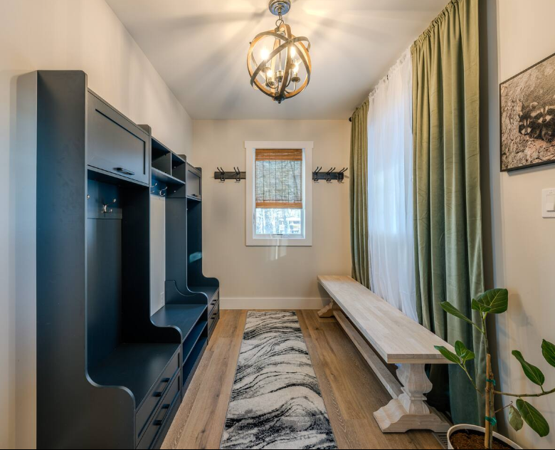 Room with window, green curtains, bench, black shelving unit, and hanging light fixture, with a rug on wooden floor.