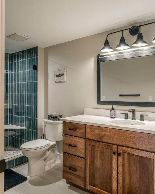 Bathroom with wooden vanity, large mirror, light fixture, toilet, and shower with green tiles.