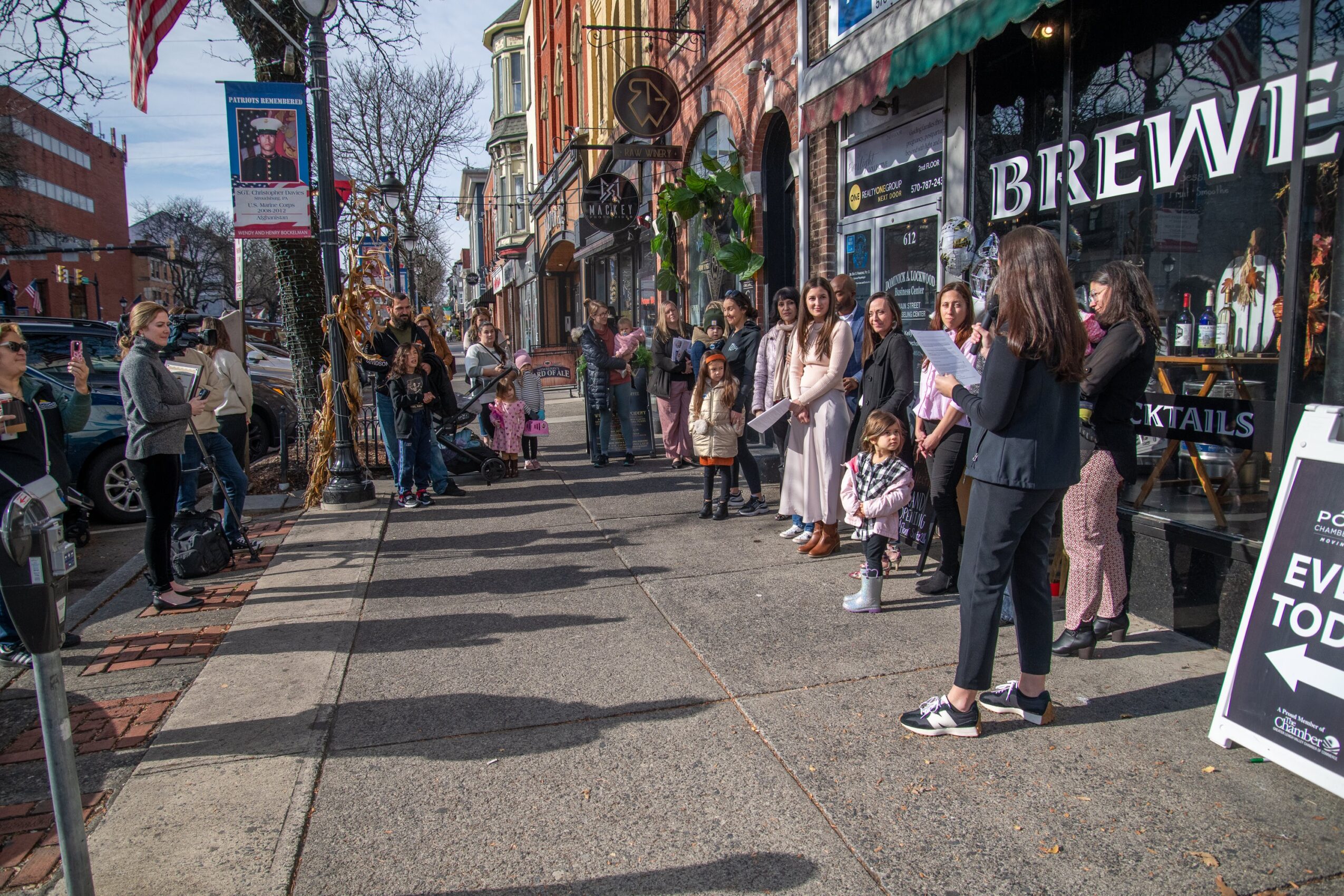 People walk along a busy city street with shops and storefronts, some standing and talking, others walking or waiting in line.