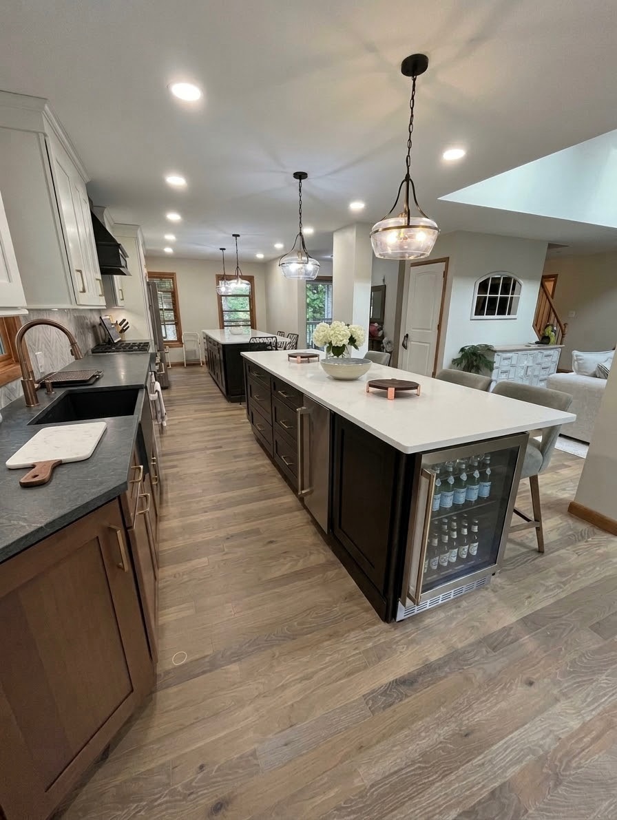 Kitchen with island, pendant lights, white countertops, dark cabinets, and a view into living area with sofa and windows.