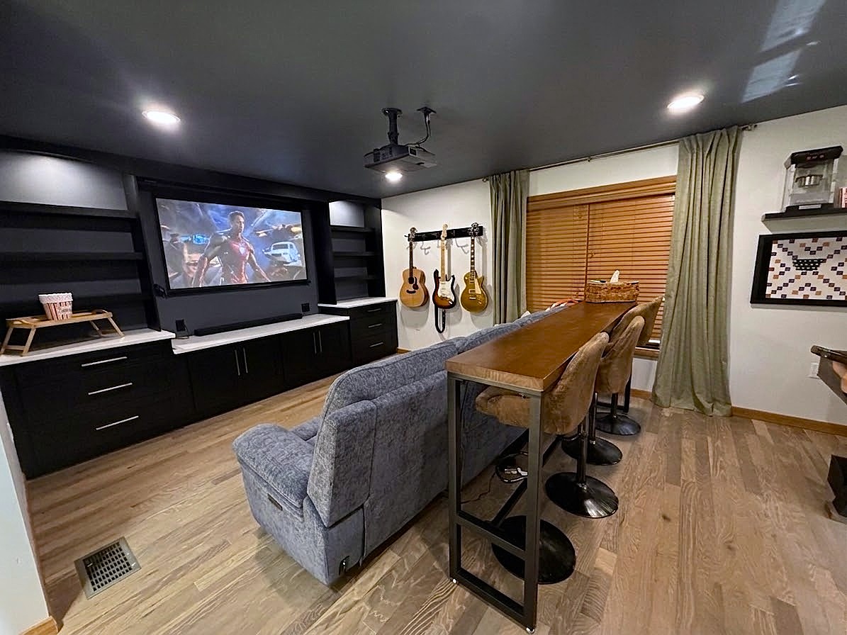 Living room with a large TV, gray sofa, wooden dining table, and musical instruments hanging on the wall.