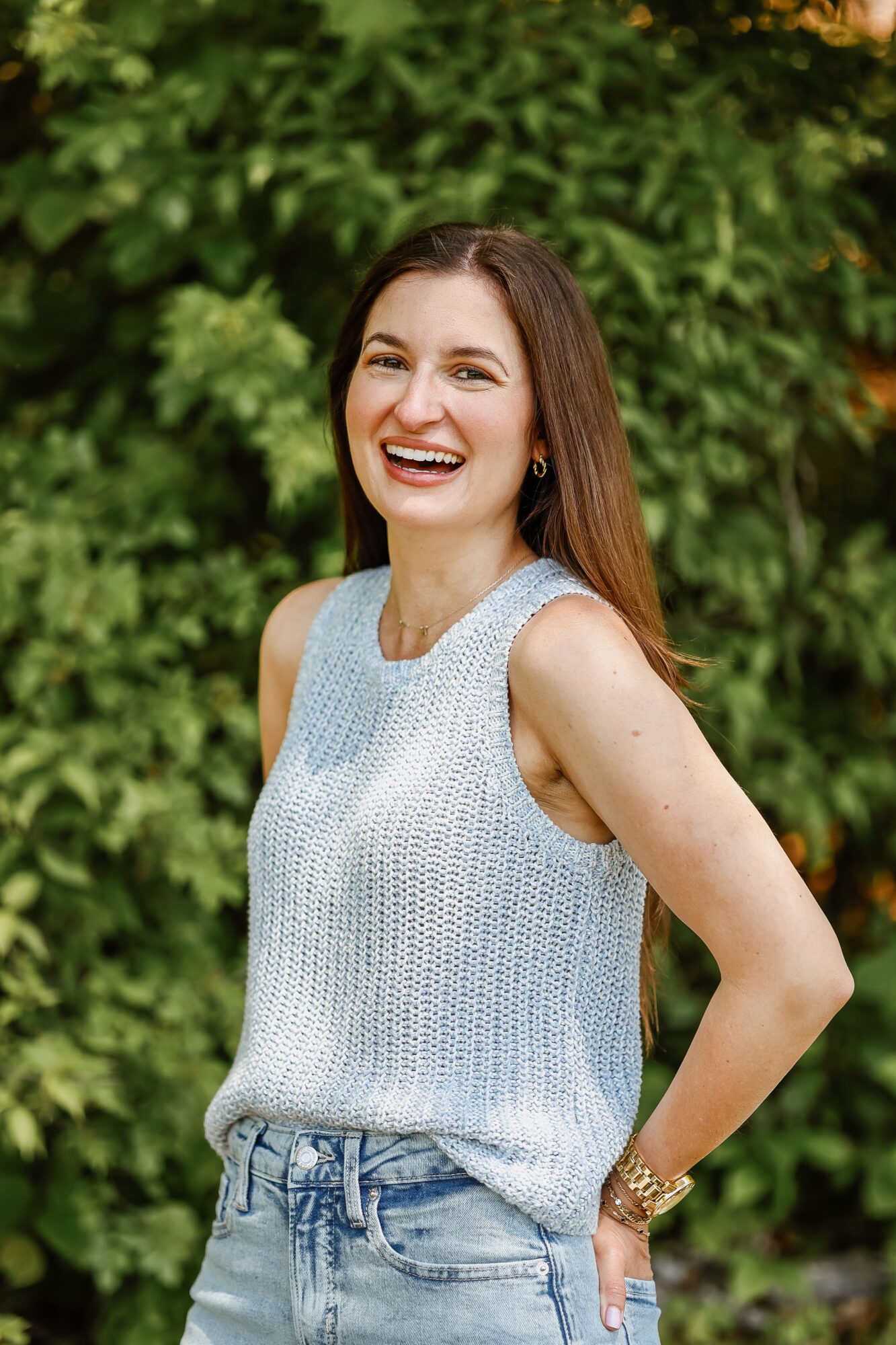 Young woman with long brown hair smiling outdoors, wearing a sleeveless light gray top and jeans, standing in front of green foliage.
