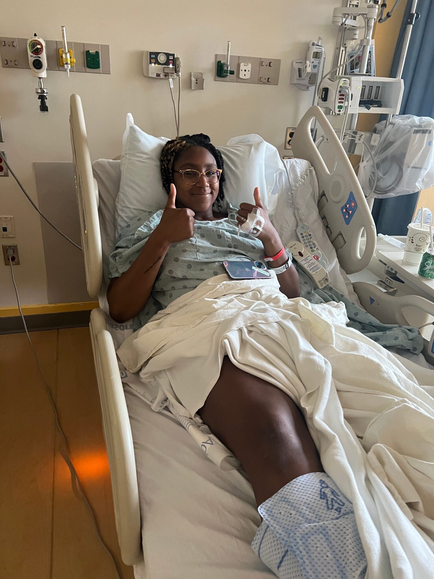 Person lying in hospital bed with medical equipment around, holding a phone, wearing hospital gown and wristbands.
