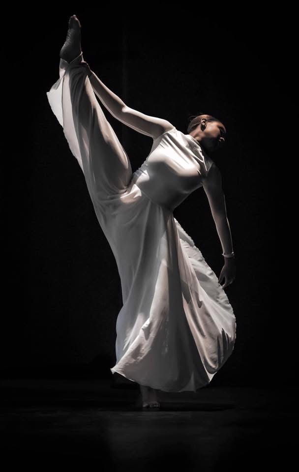 A dancer in a flowing white dress performs a high leg lift against a dark background.