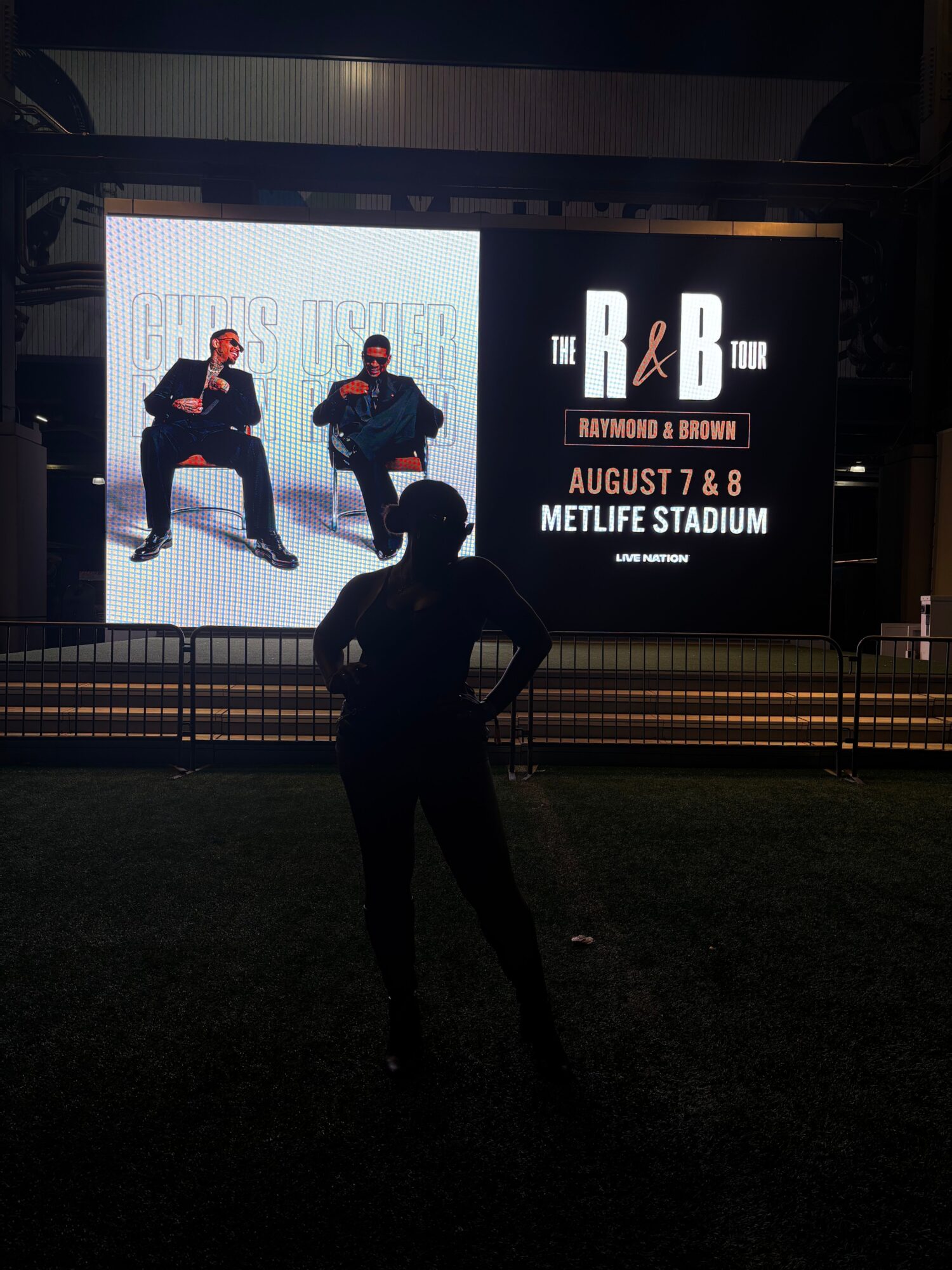 Two people in dark clothing and red hats pose in front of a large illuminated sign at night, with a fence and grass nearby.