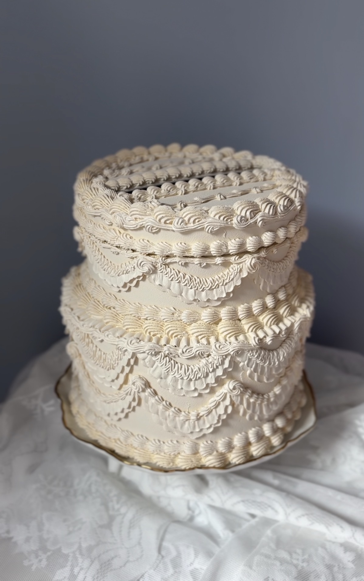 Three-tiered cake with intricate white icing decorations, placed on a white cloth against a dark background.