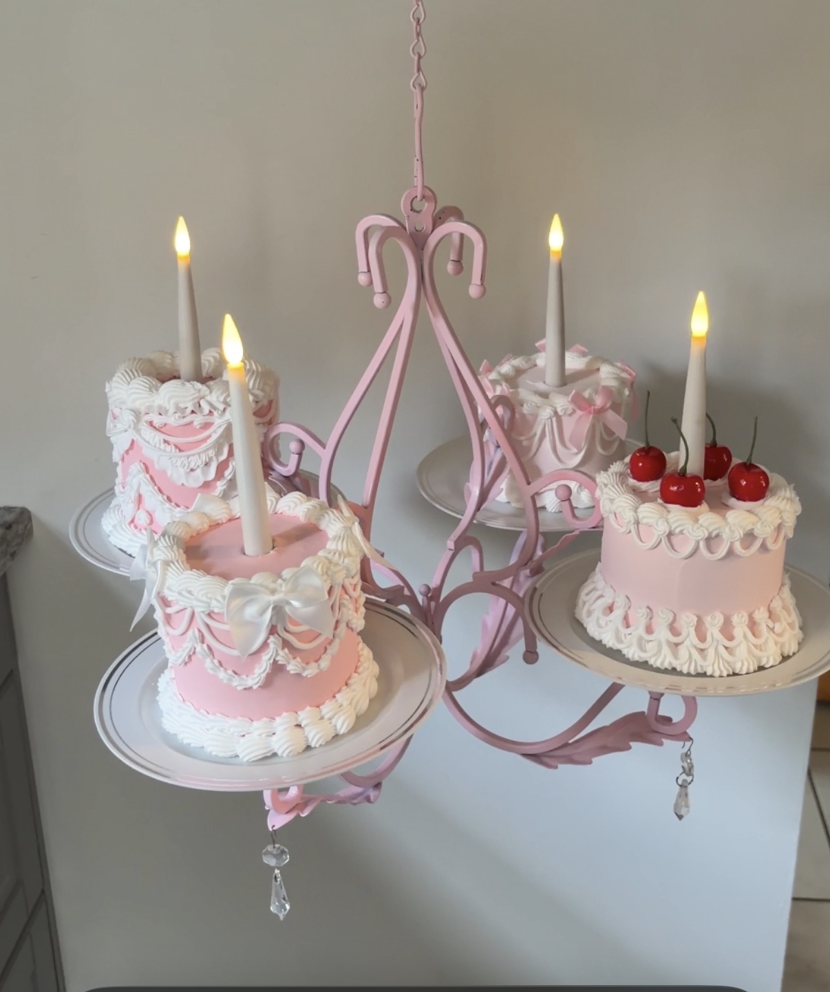 Three pink birthday cakes with lit candles and cherries, decorated with white icing and pink ribbons, on a pink chandelier stand.