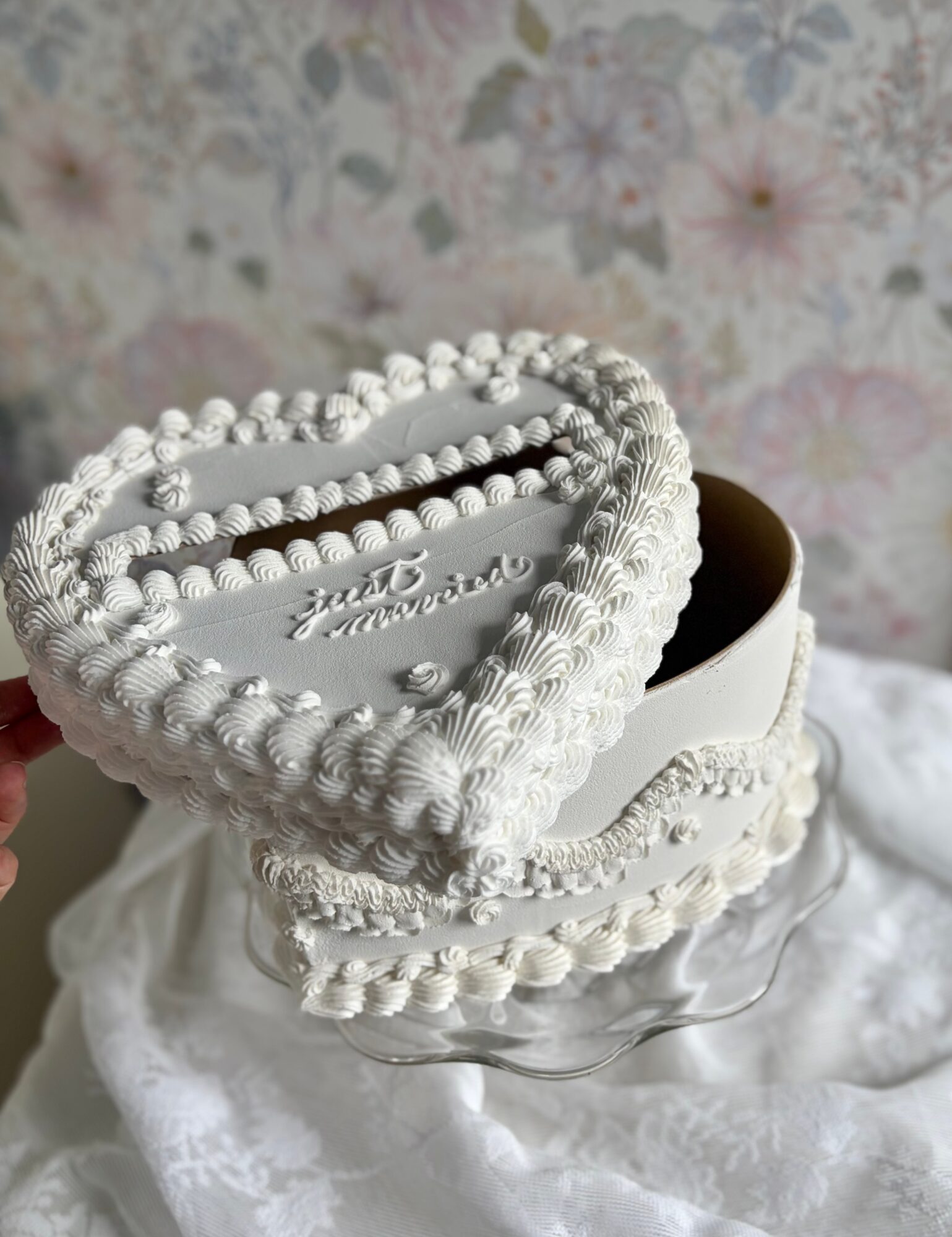 Heart-shaped decorated box with white icing and beads, on a white cloth with floral wallpaper background.