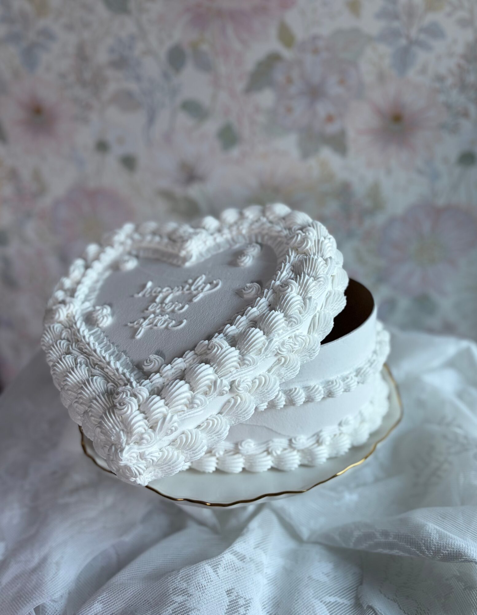 Heart-shaped cake with white frosting and decorative piping, on a white cloth, with a floral background.