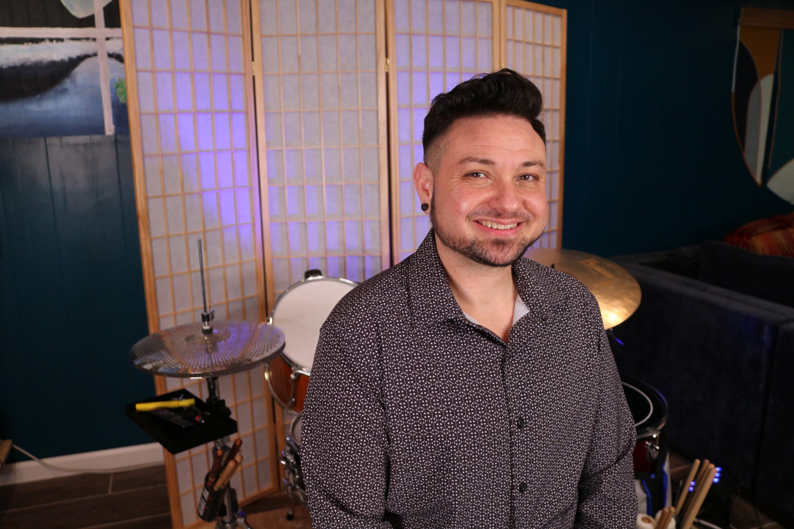 Smiling man with dark hair and beard, wearing a patterned shirt, standing indoors with a folding screen behind him.