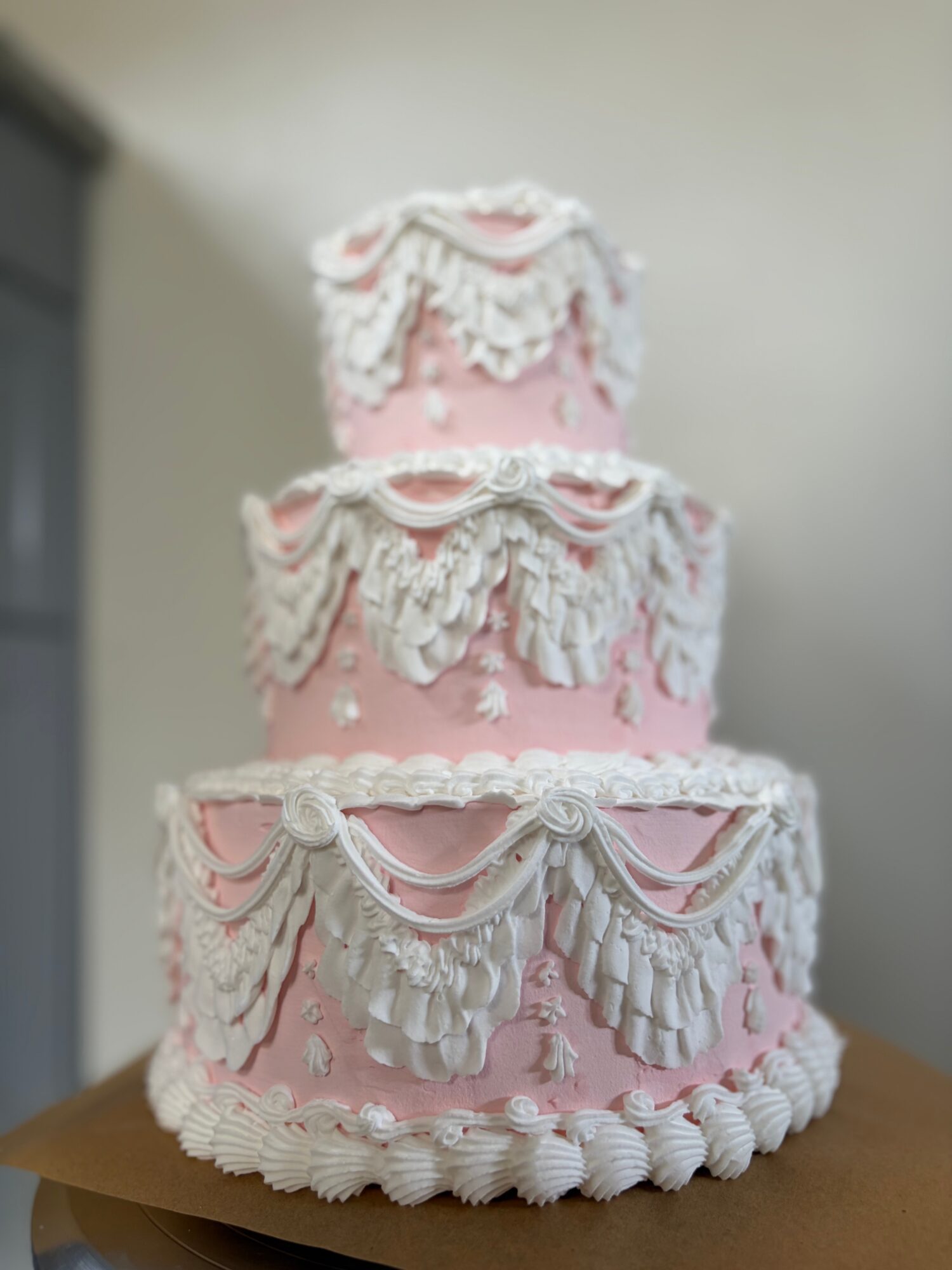 Three-tier pink cake decorated with white icing in ornate, draped patterns, on a wooden surface.