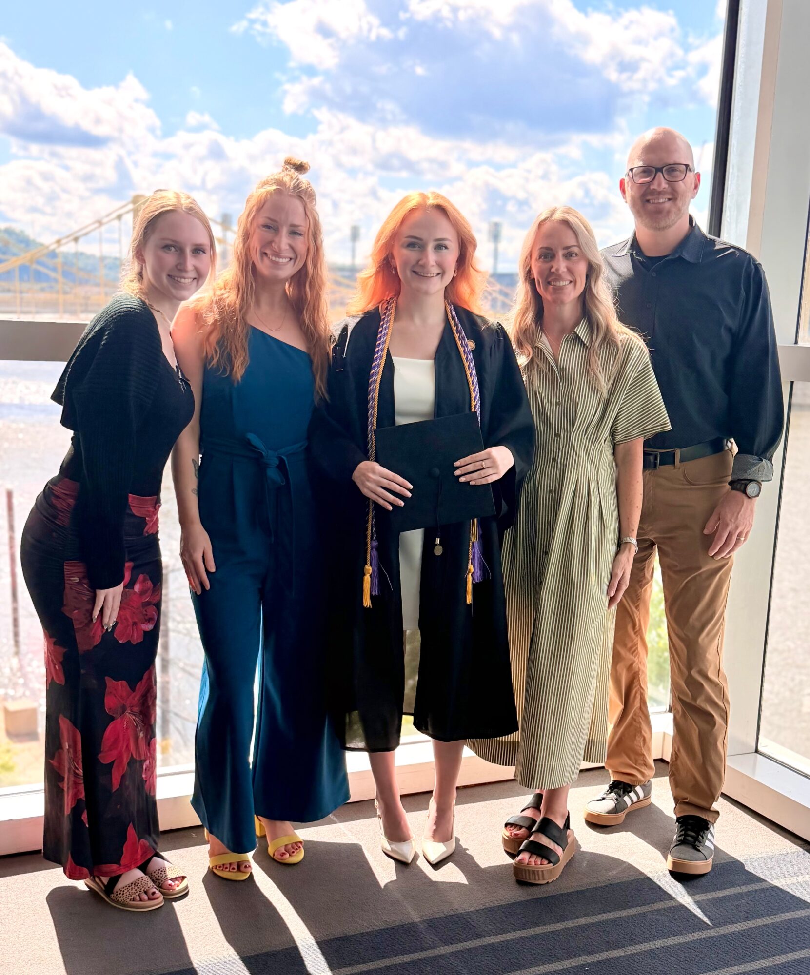 Group of five people standing indoors near large window with sky and bridge view, smiling, dressed casually. 20 words.