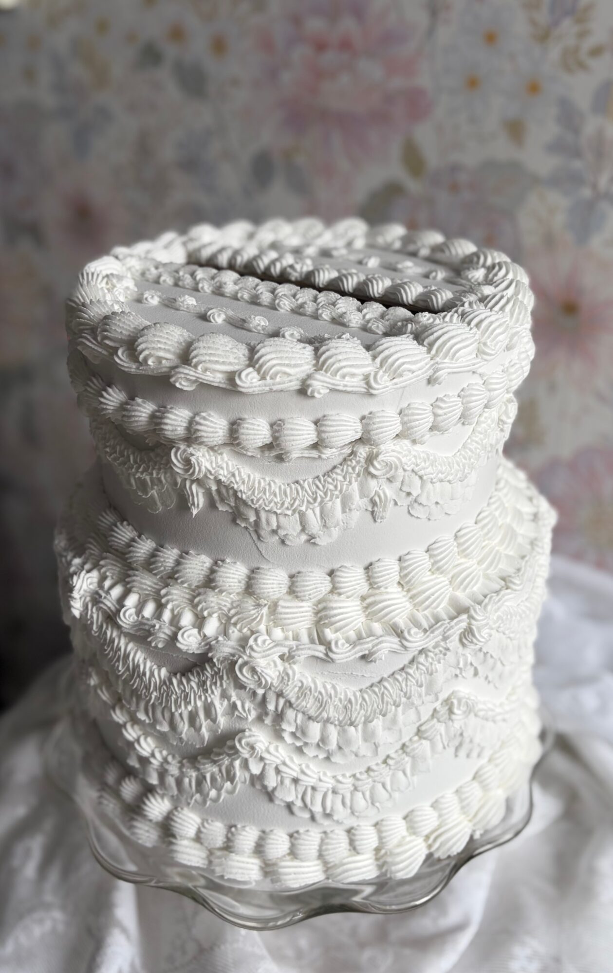 Stack of decorated white cakes with intricate piping, placed on a glass cake stand, against a floral background.