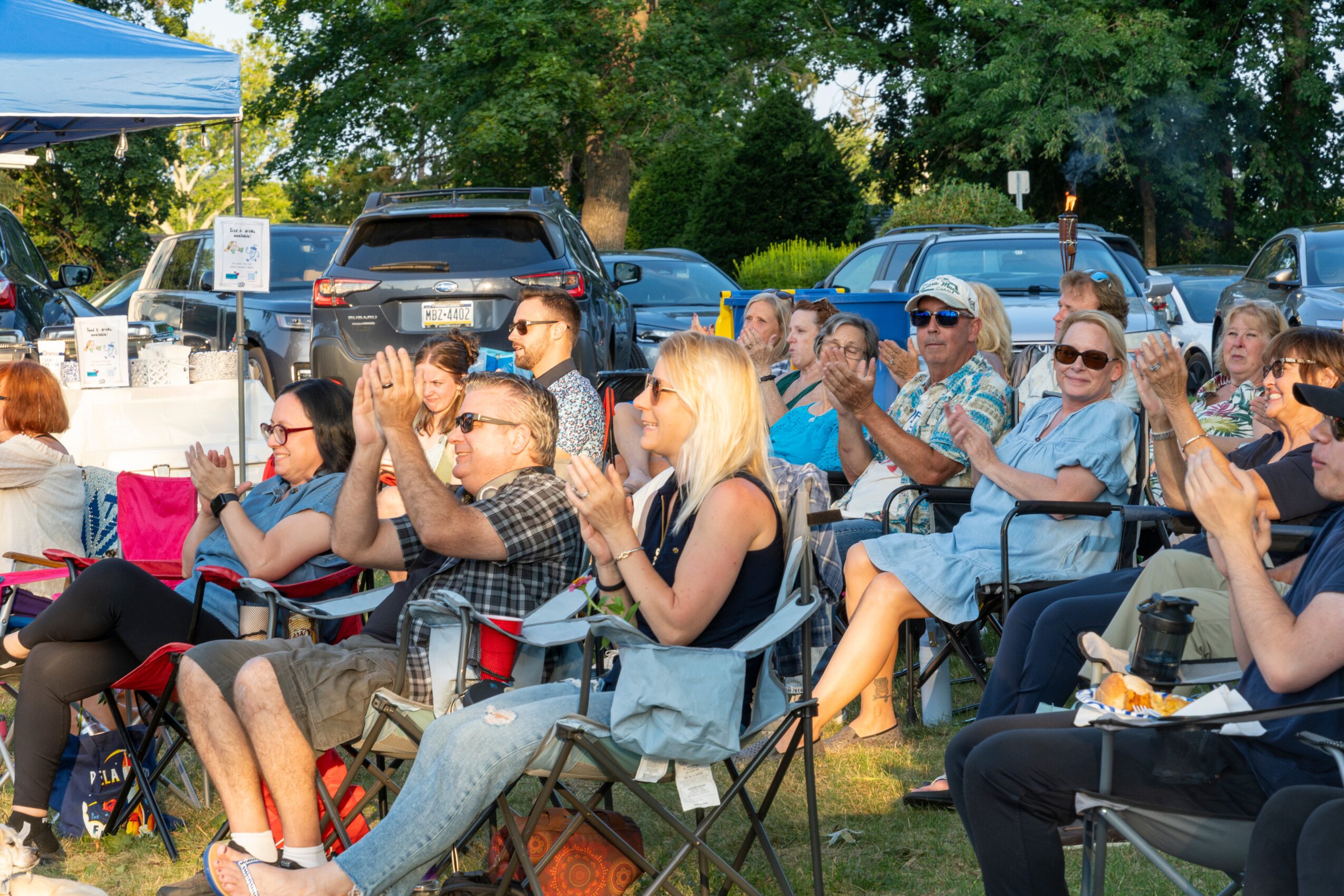 Group of people sitting outdoors, clapping and enjoying an event, with trees and parked cars in the background.