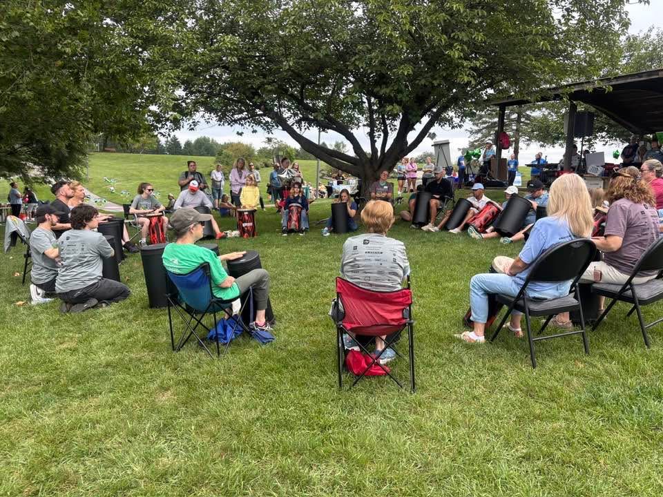 Group of people gathered outdoors on grass, sitting on chairs and standing under a tree, with a stage and trees in background.