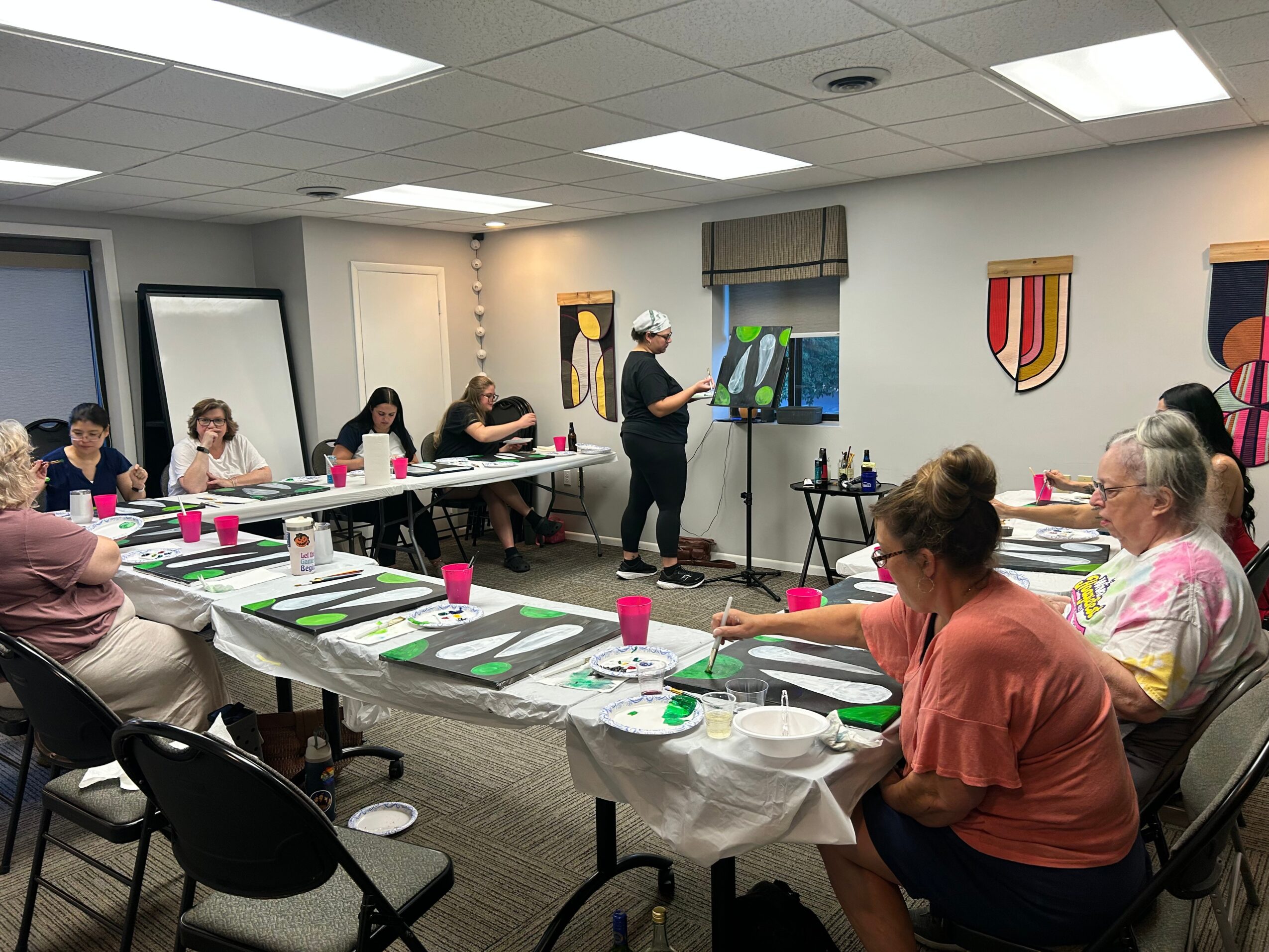 People seated around a table in a classroom, listening to a woman presenting artwork, with colorful paintings on the wall.
