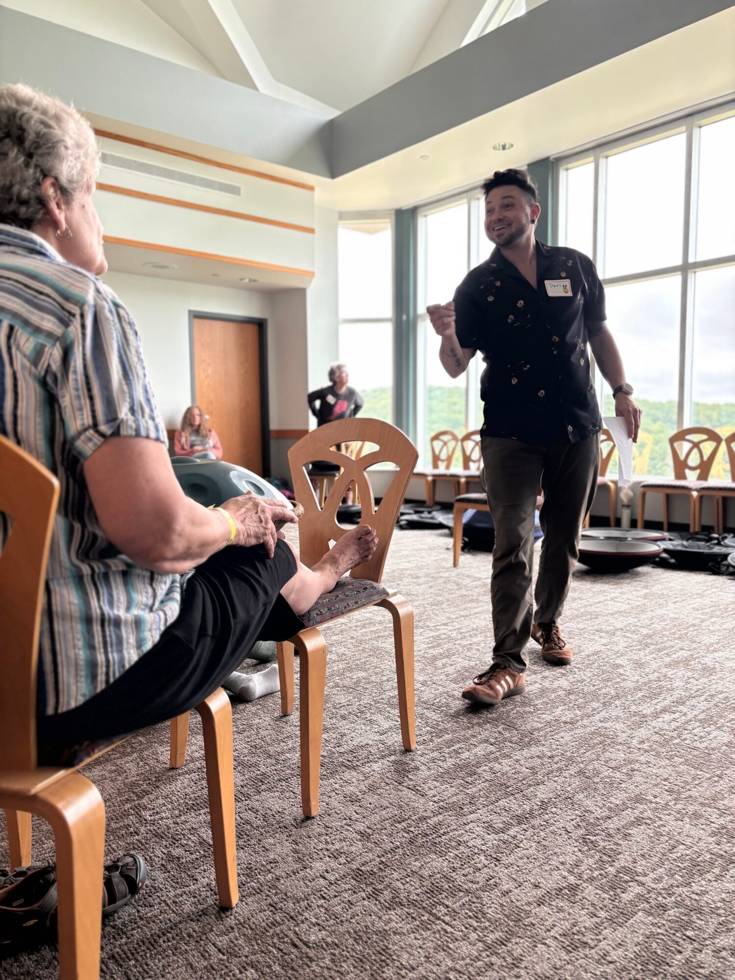 Person standing and speaking to seated person in a bright room with large windows and chairs.
