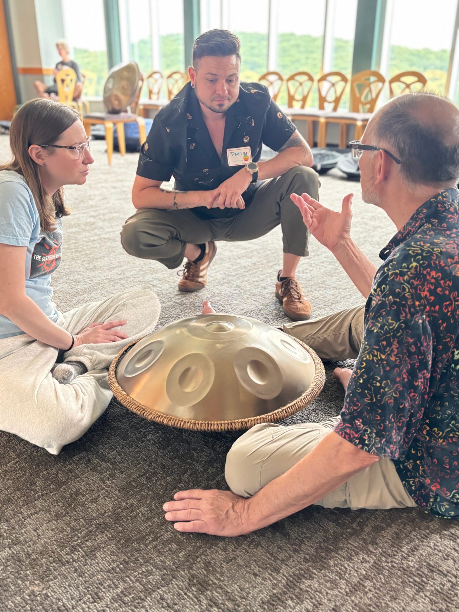 Three people sit on the floor around a handpan instrument, engaging in a discussion indoors with chairs and windows in the background.
