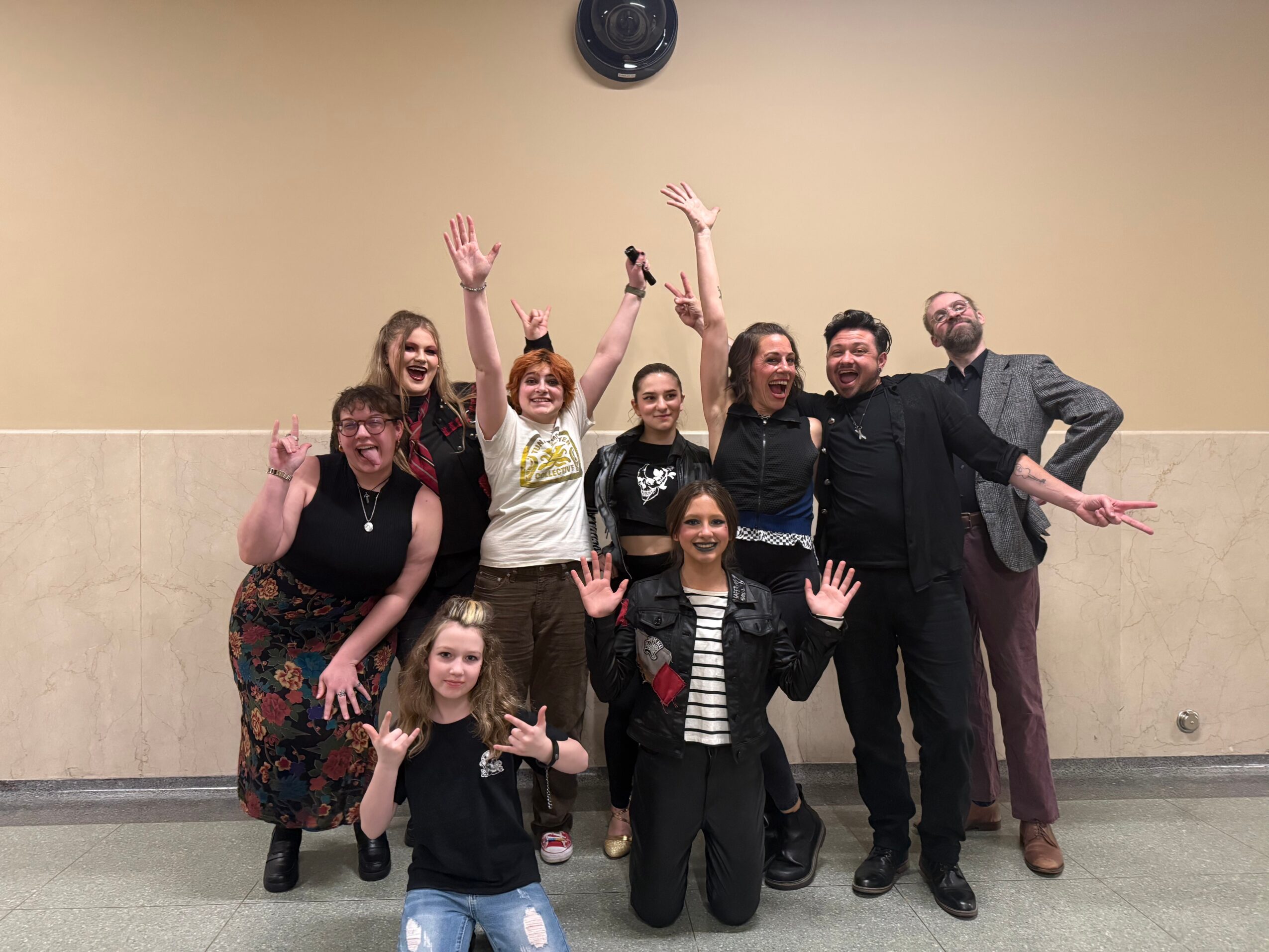 Group of nine people posing and smiling in a room with a beige wall, some raising hands, some kneeling or standing.