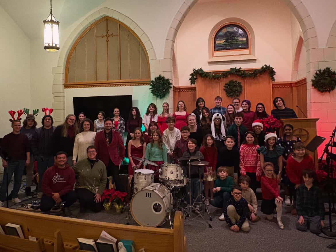 Group of people gathered in a church, some wearing holiday accessories, with musical instruments and festive decorations.