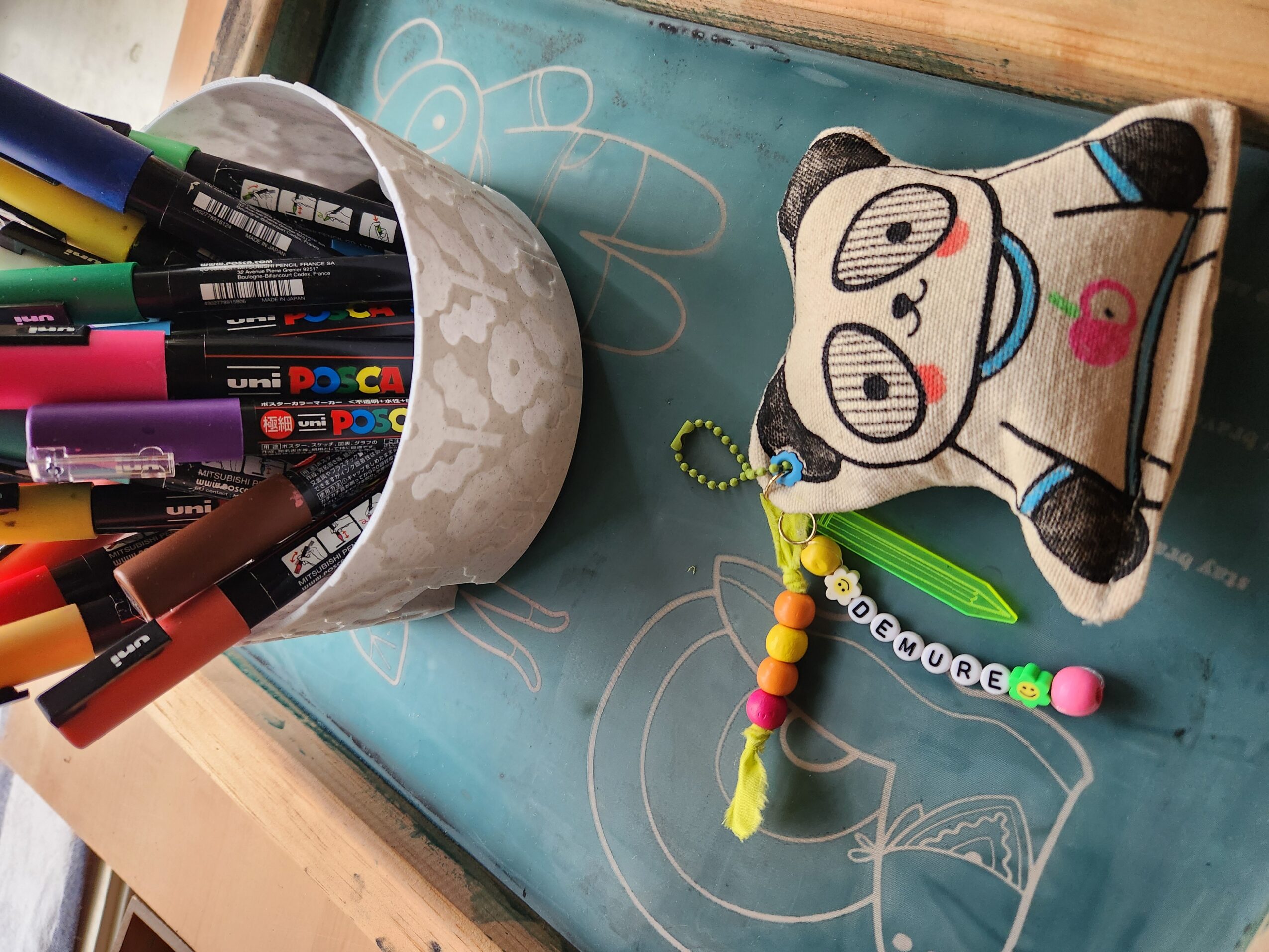 A pencil holder filled with colorful markers, a plush toy with glasses, and a beaded keychain on a blue chalkboard surface.
