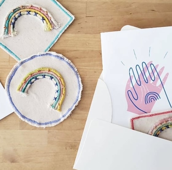 Two embroidered rainbow patches on white fabric, next to a paper with a hand illustration, on a wooden surface.