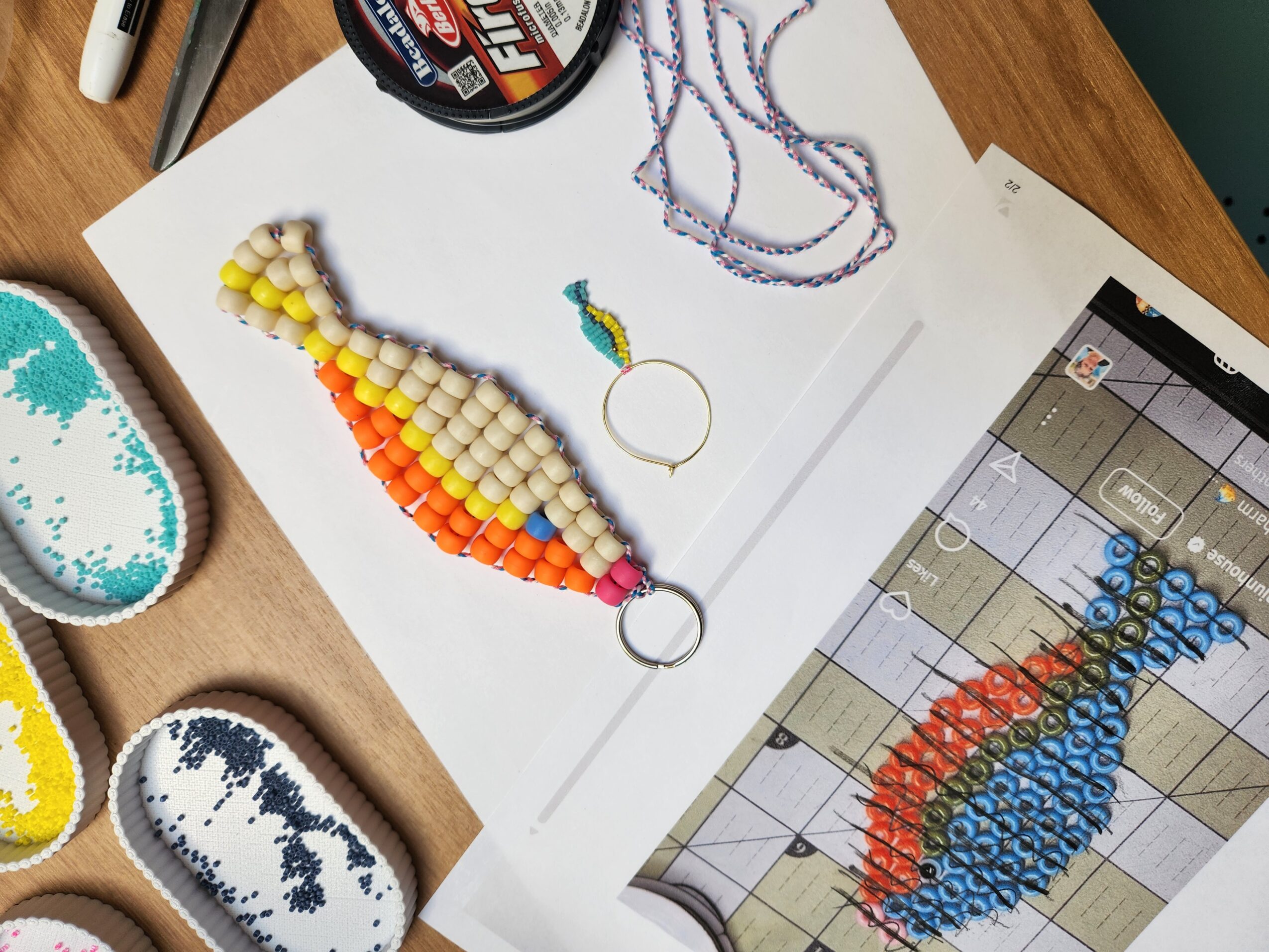 Paper with a colorful beaded fish, a rainbow-colored beaded fish, and a beaded fish pattern on a tablet screen, on a wooden table.